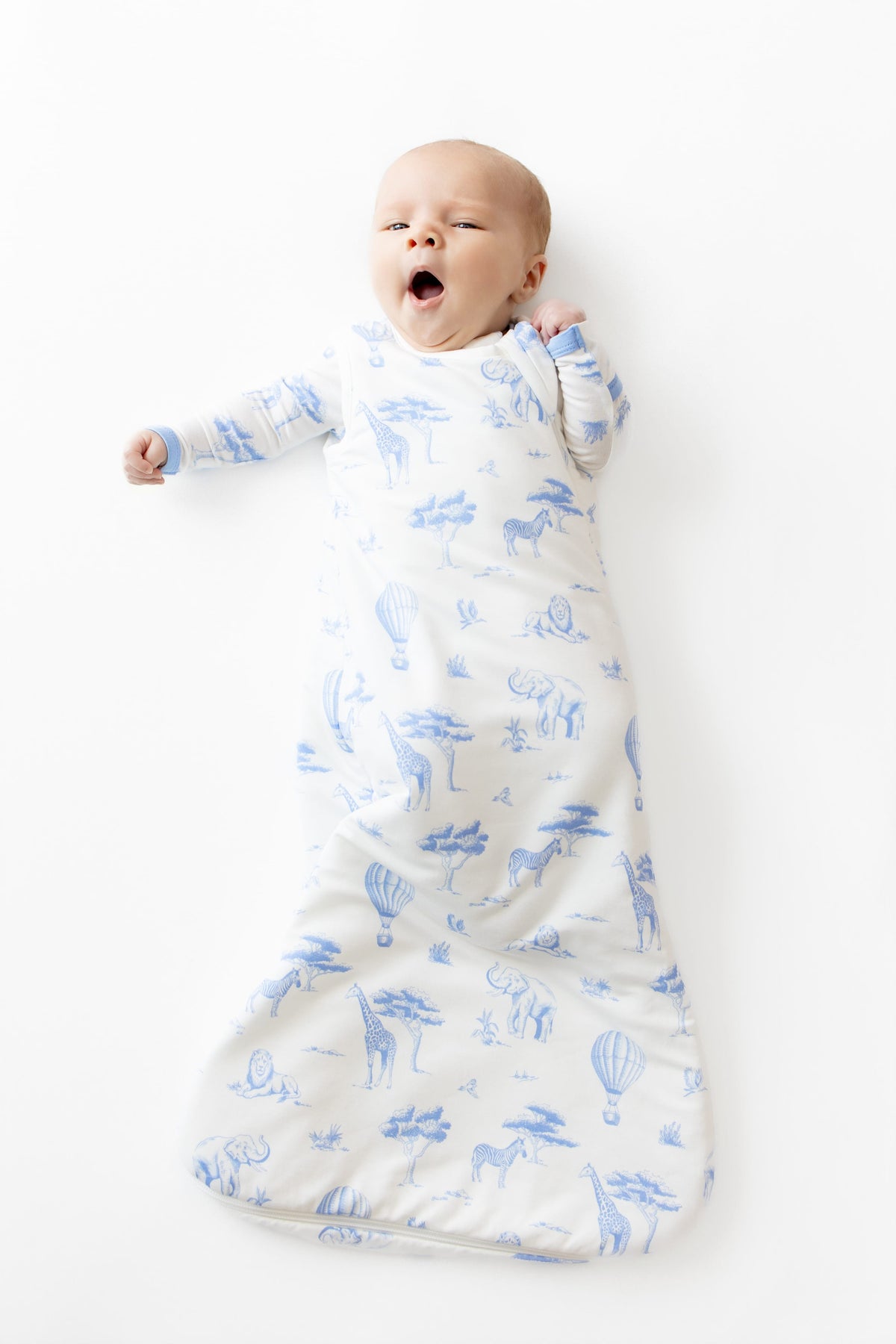 Yawning infant laying on a white surface wearing the Sleep Bag in Toile Safari 1.0 and matching romper