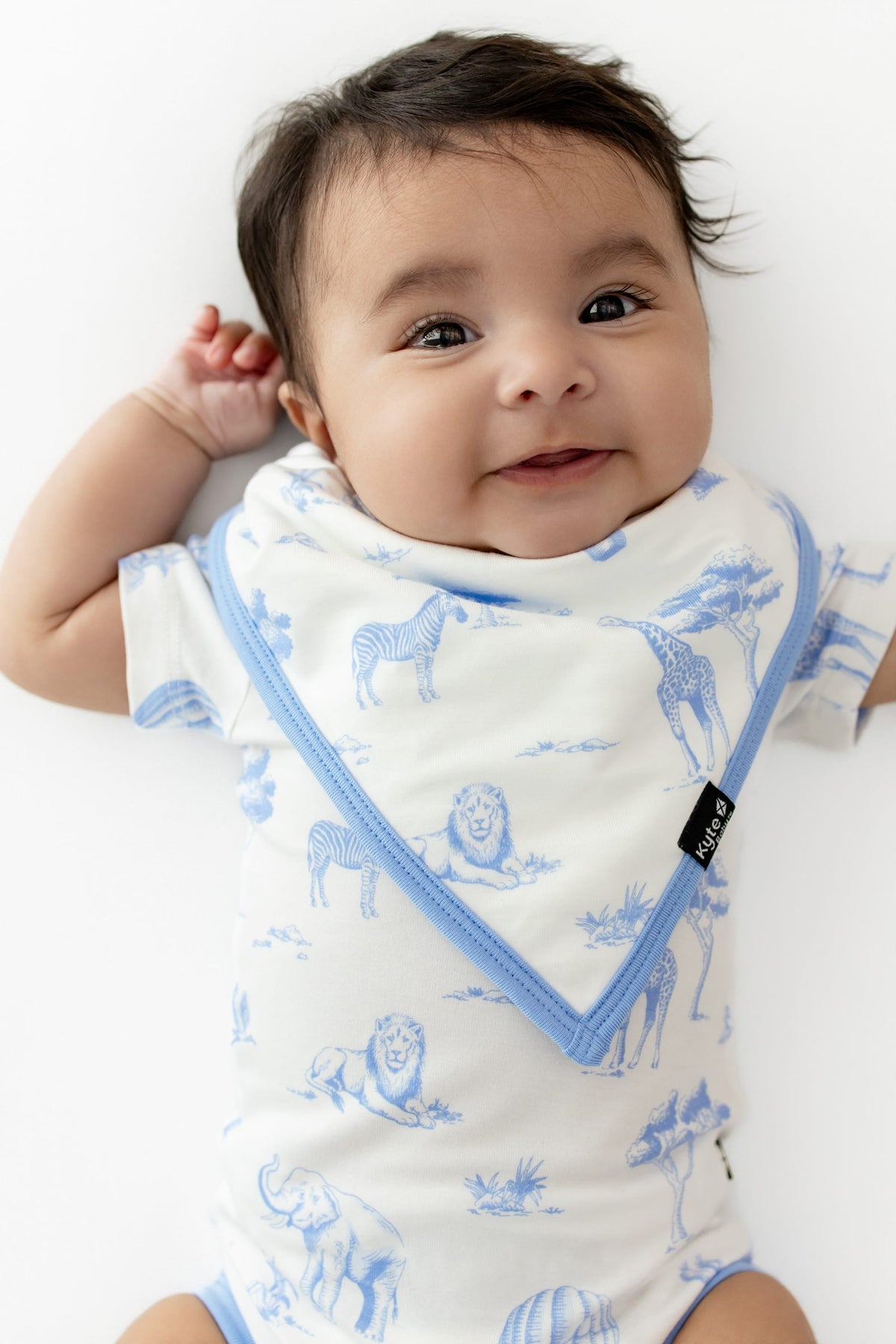Infant laying on a white surface wearing the Bib in Toile Safari with a matching bodysuit