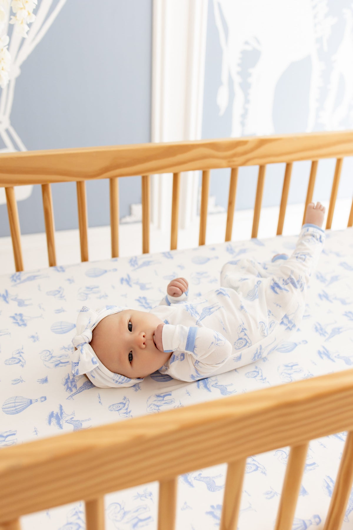 Infant laying in a light wooden crib on top of a Crib Sheet in Toile Safari