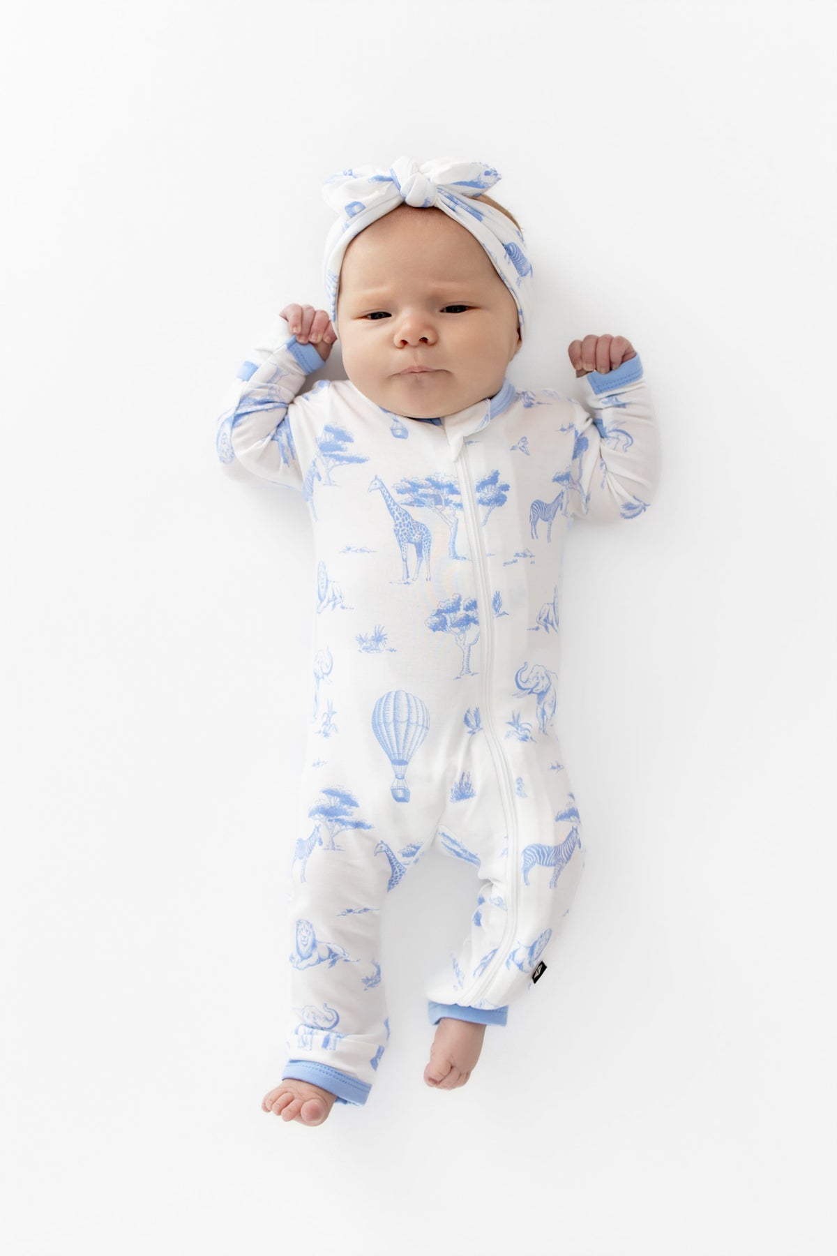 Newborn girl laying on a white surface wearing the Zippered Romper in Toile Safari and matching headband