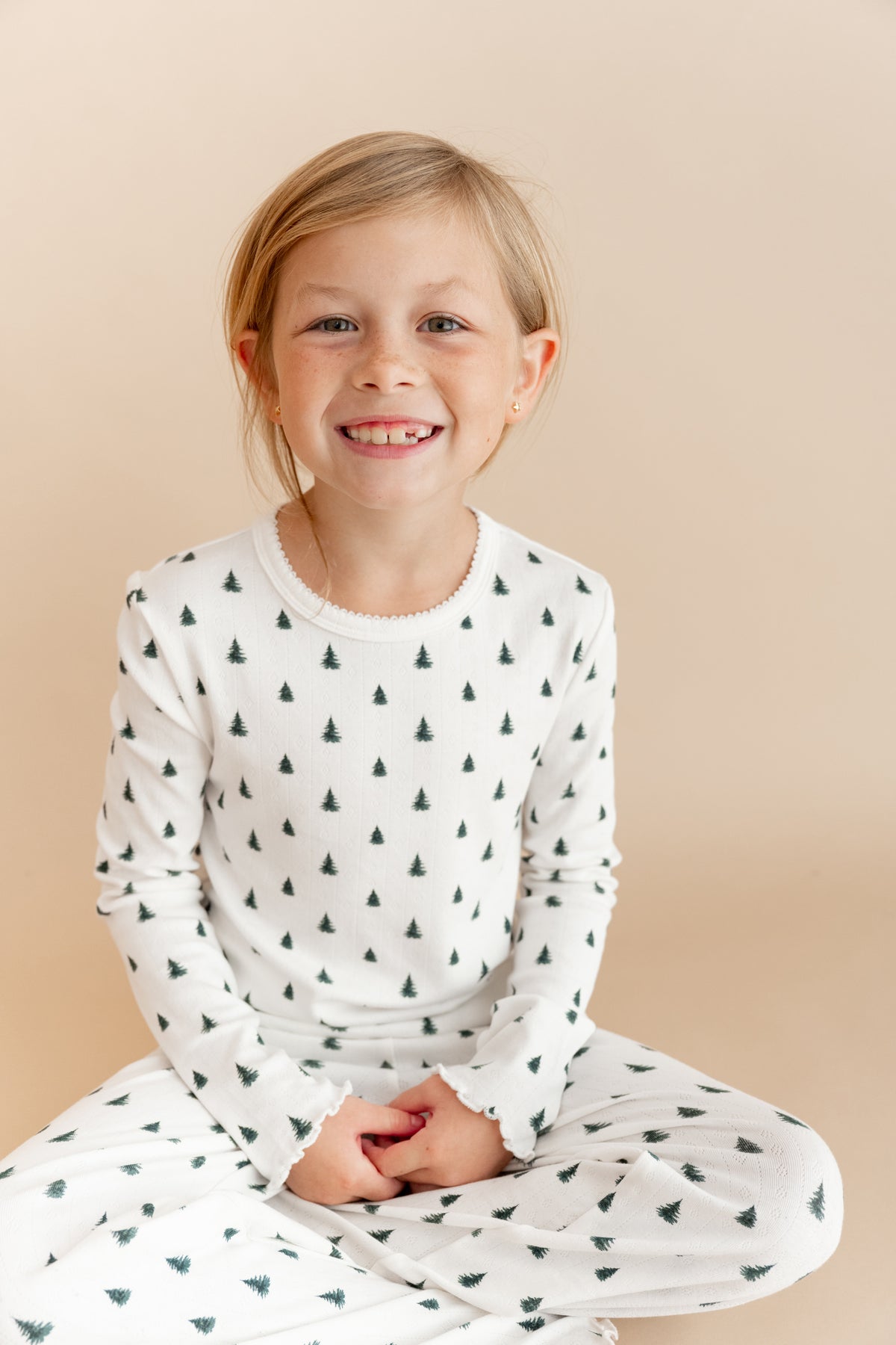 A young girl with blonde hair sitting cross-legged, wearing a white long-sleeve pointelle top and matching pants with a small dark green pine tree pattern, against a solid beige background.