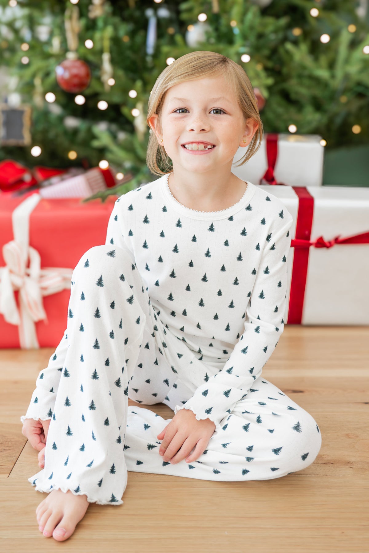 A young girl with blonde hair sits smiling on a wooden floor in front of a decorated Christmas tree and wrapped gifts. She is wearing a white long-sleeve pointelle set with a repeating pattern of small dark green pine trees.