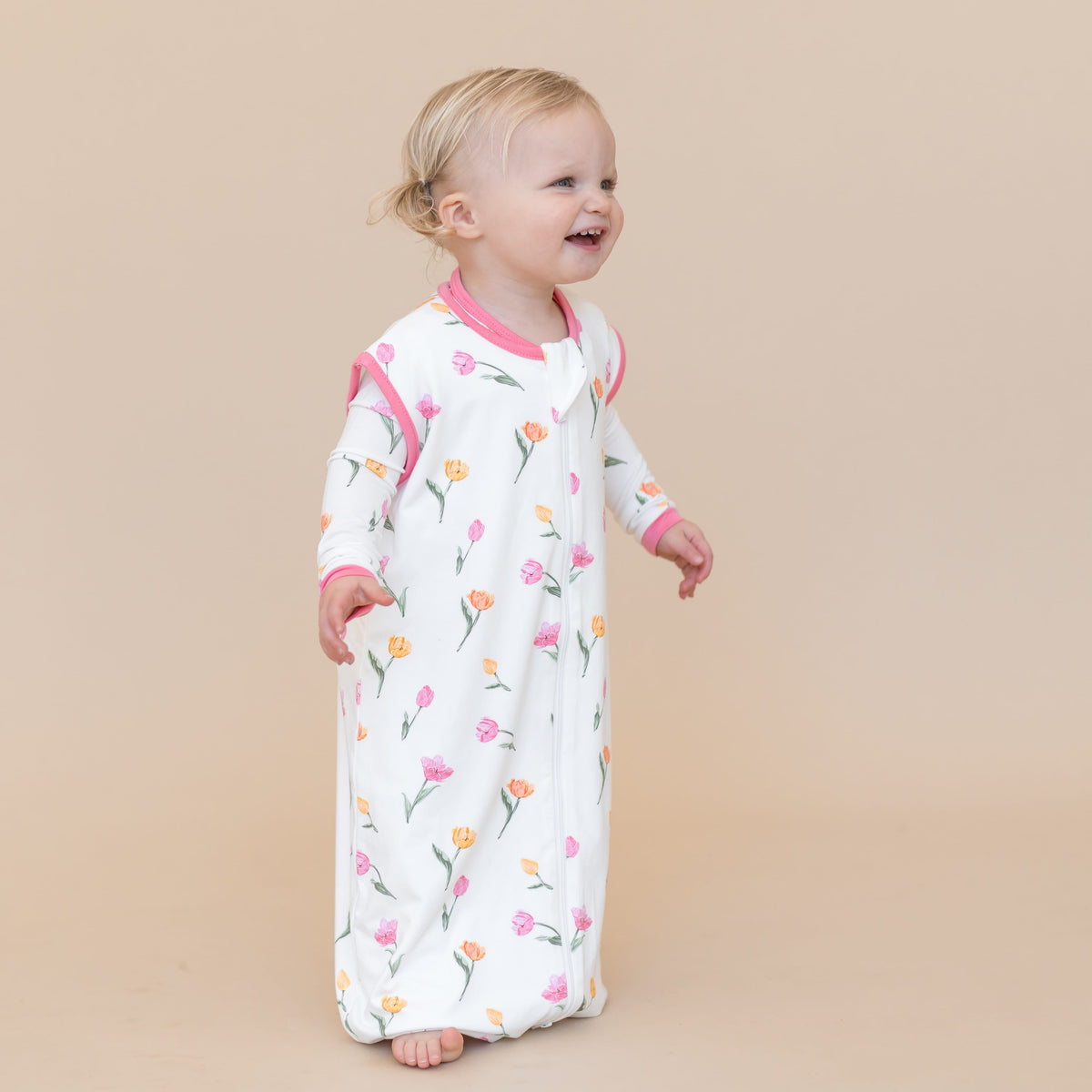 Young smiling toddler standing wearing the Sleep Bag Walker in Tulip 0.5