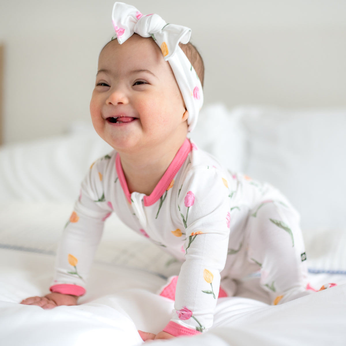 Smiling toddler sitting on a bed wearing the Zippered Footie in Tulip and matching bow