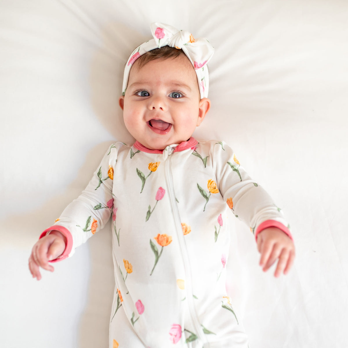 Close up of a toddler laying on a bed wearing the Zippered Romper in Tulip with matching knotted bow headband