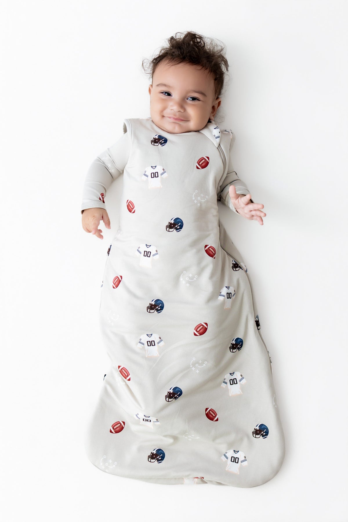 A baby lying on their back wearing a long-sleeve sleep sack with a white base and a pattern of American footballs and football helmets in red, blue, and white, on a solid white background.