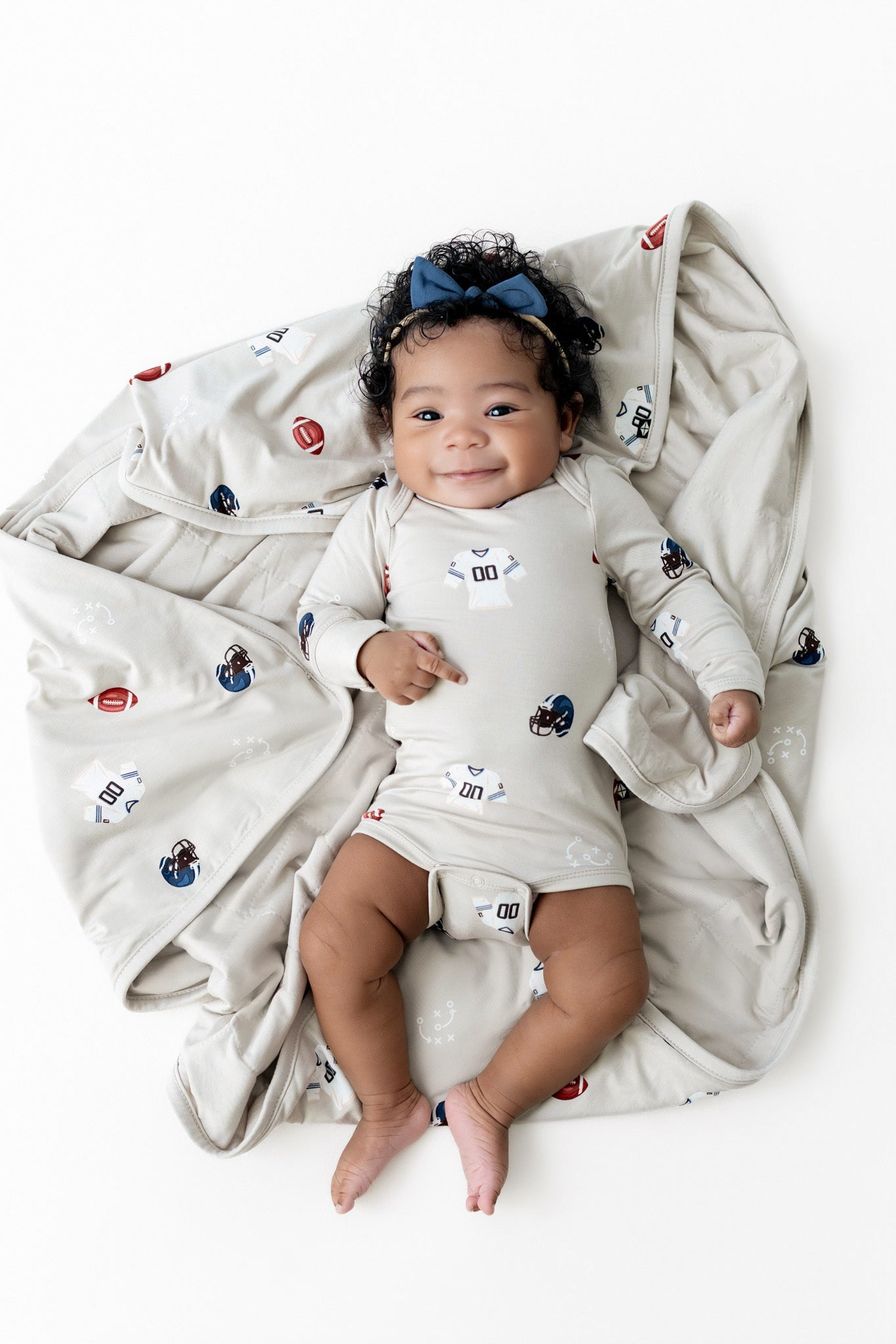 A baby lying on a light beige blanket with a football-themed print, wearing a matching long-sleeve bodysuit and a blue bow headband, photographed from above on a white background.