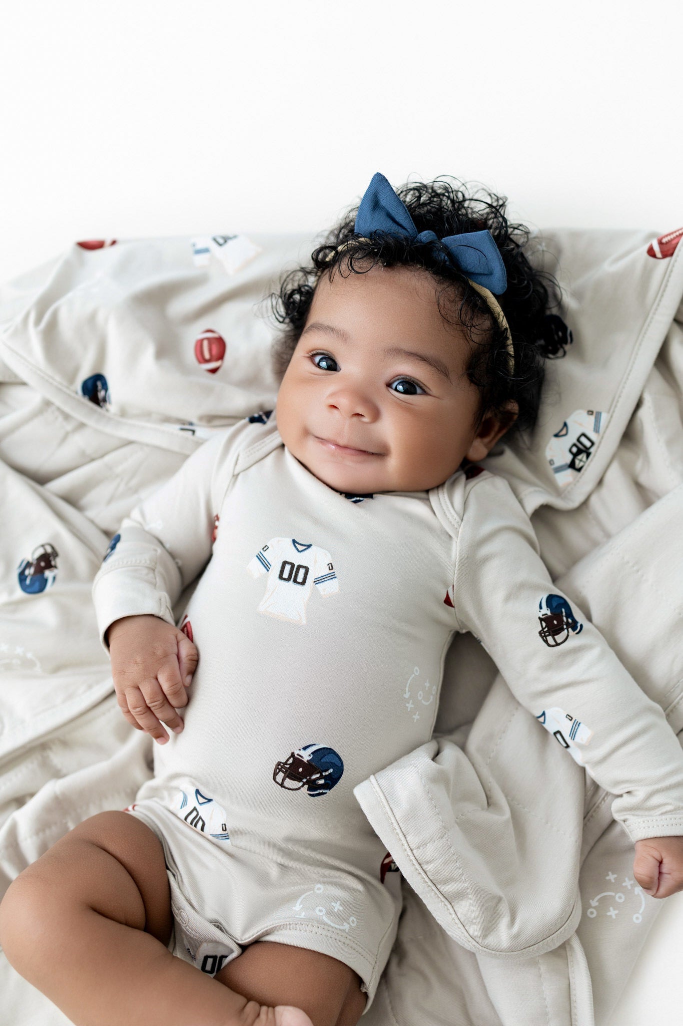 A smiling baby lying on a white blanket, wearing a long-sleeve cream bodysuit with football team graphics and a blue bow headband.