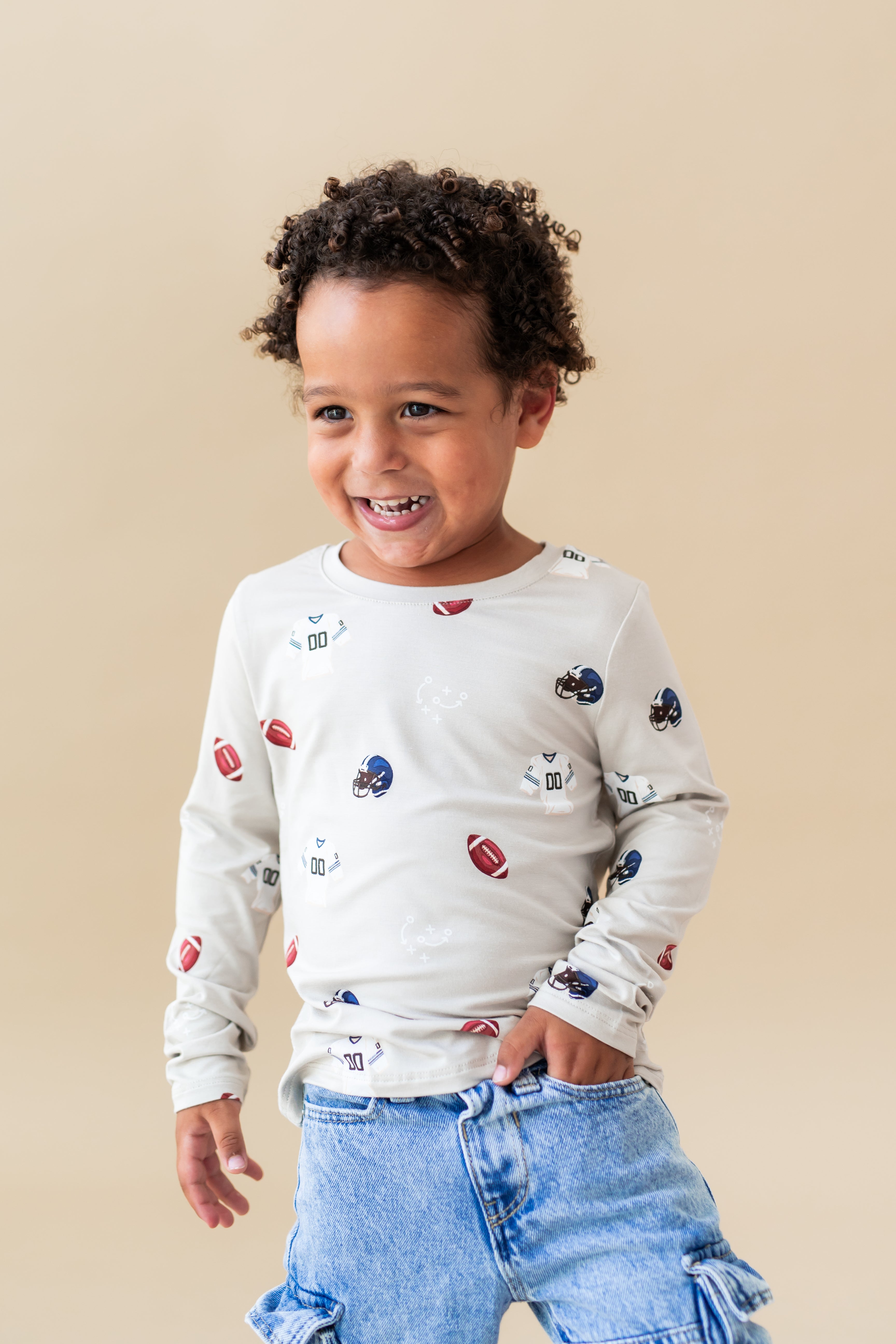 Boy wearing Long Sleeve Toddler Crew Neck Tee in Vintage Football print over blue jeans on a beige background