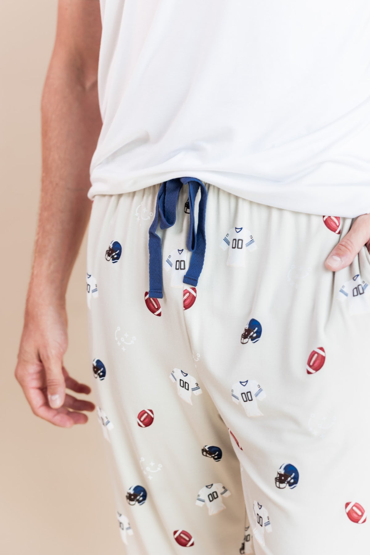 Close-up of a person wearing cream-colored athletic pajama pants with a blue drawstring and an all-over print of football helmets and footballs, paired with a white tank top.