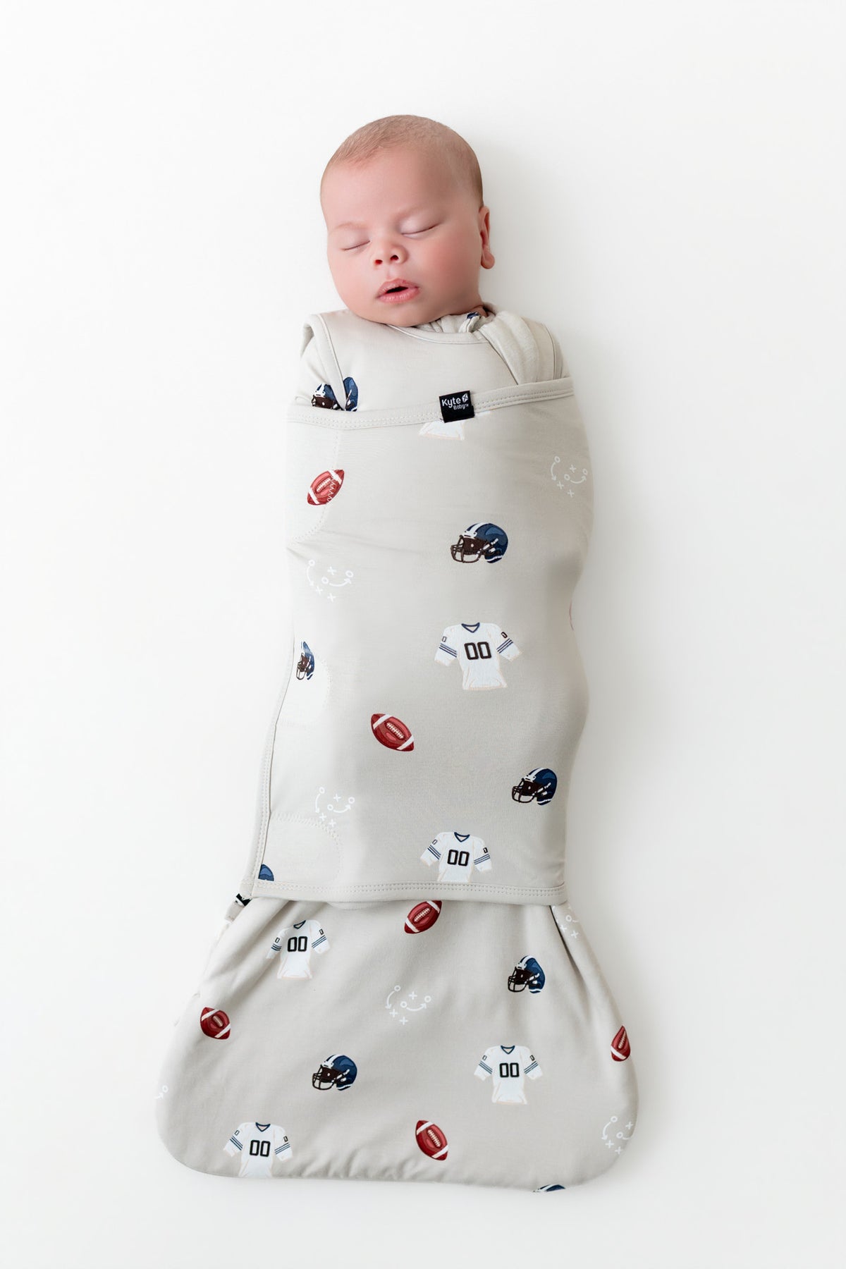A sleeping newborn baby is swaddled in a light gray wrap blanket featuring a pattern of footballs, helmets, and sports jerseys, laid flat on a white background.