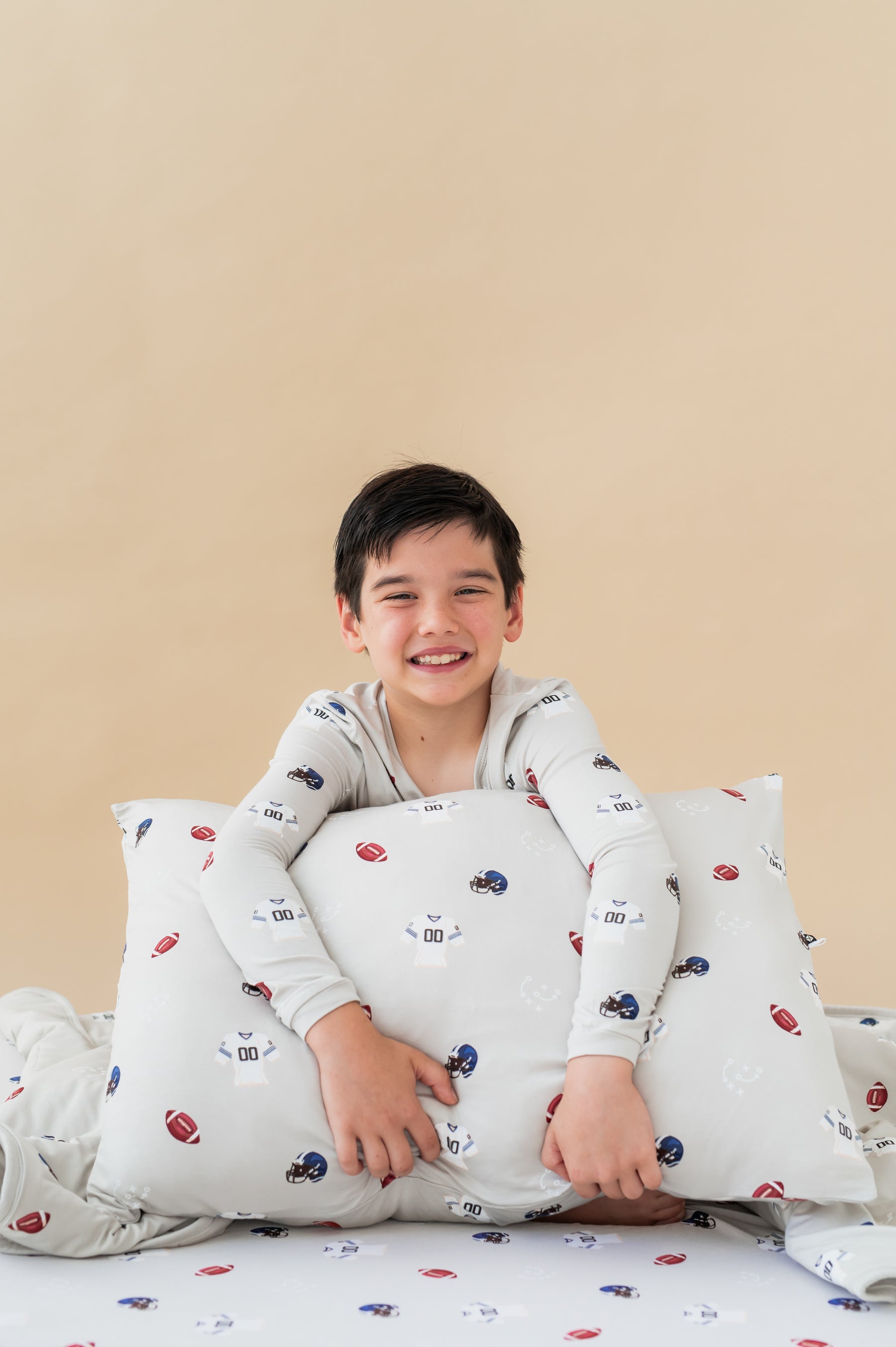 Boy holding Standard Pillowcase in Vintage Football print sitting on a similar print sheets, wearing a similar print Long sleeve pajama set on a beige background