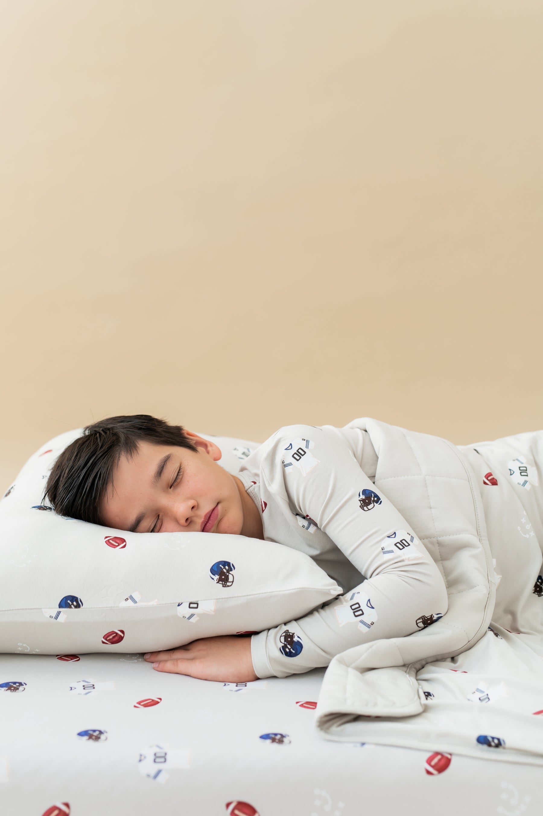 Boy sleeping on Standard Pillowcase and sheets in Vintage Football print wearing a similar print Long sleeve pajama set covered with similar print youth blanket on a beige background