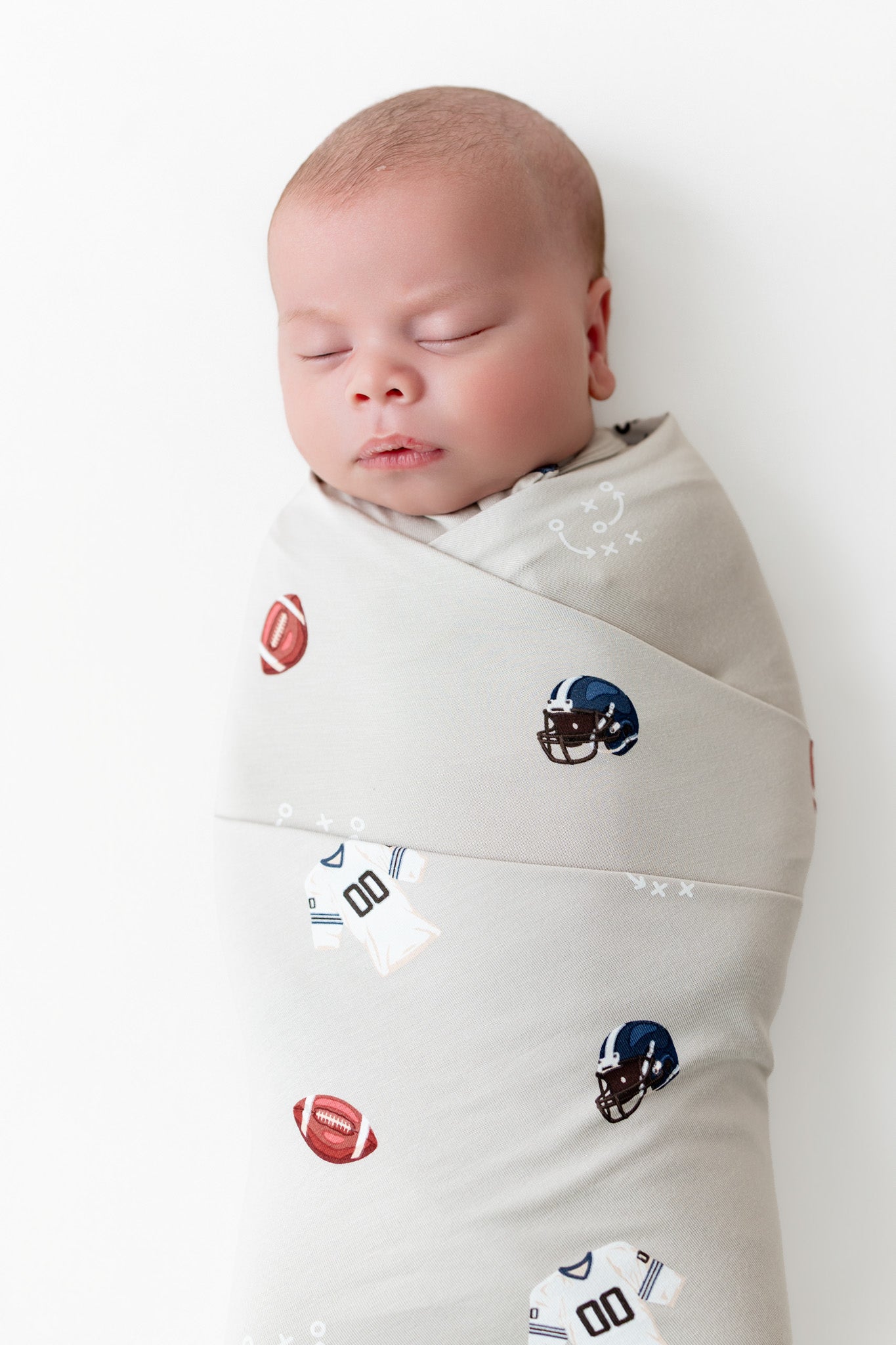 A sleeping newborn baby swaddled in a light beige fabric wrap with a pattern of footballs and football helmets, photographed against a clean white background.