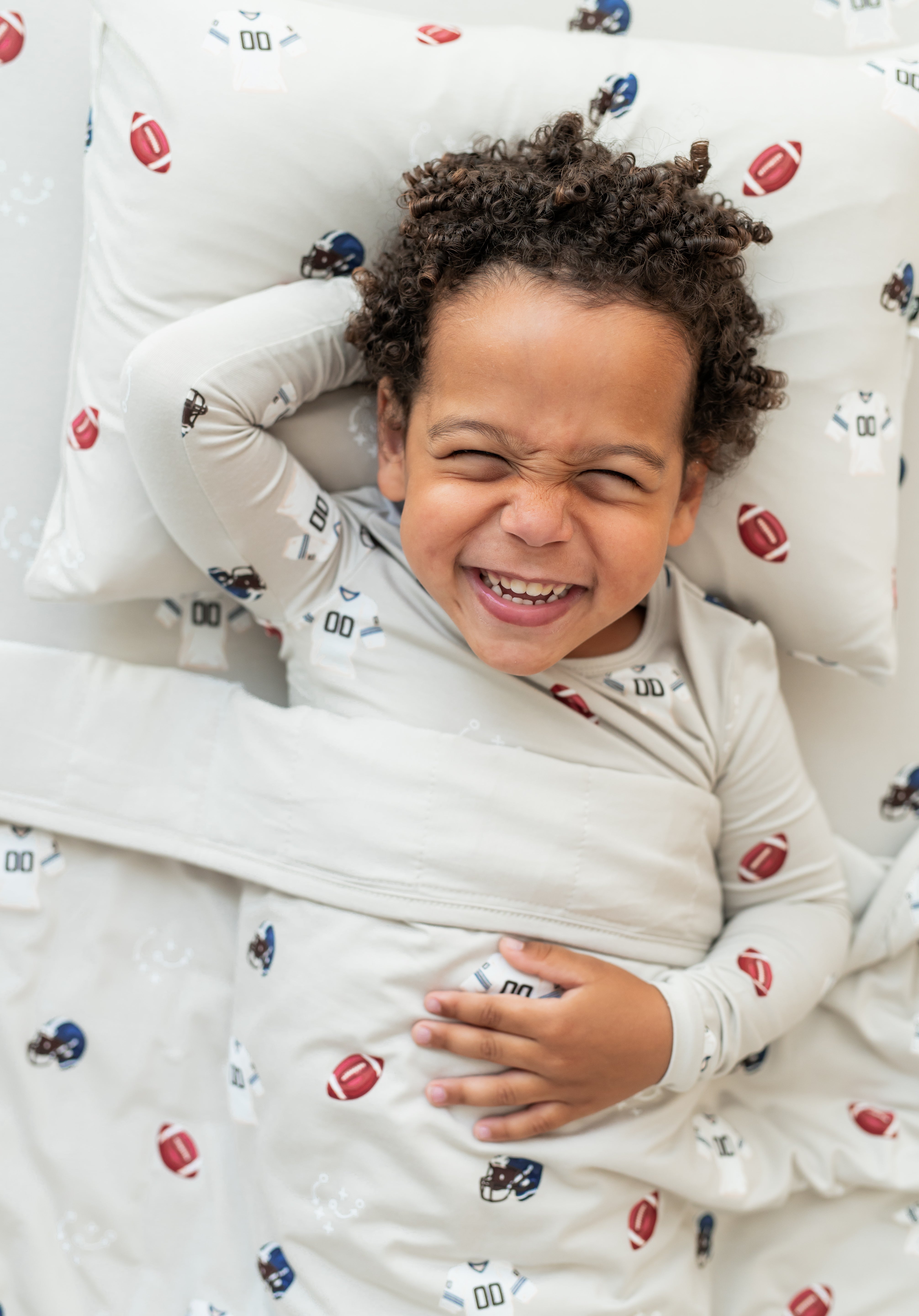 Boy lying on Toddler Pillowcase and sheet in Vintage Football print while wearing a similar print long sleeve pajama set covered with a similar print toddler blanket