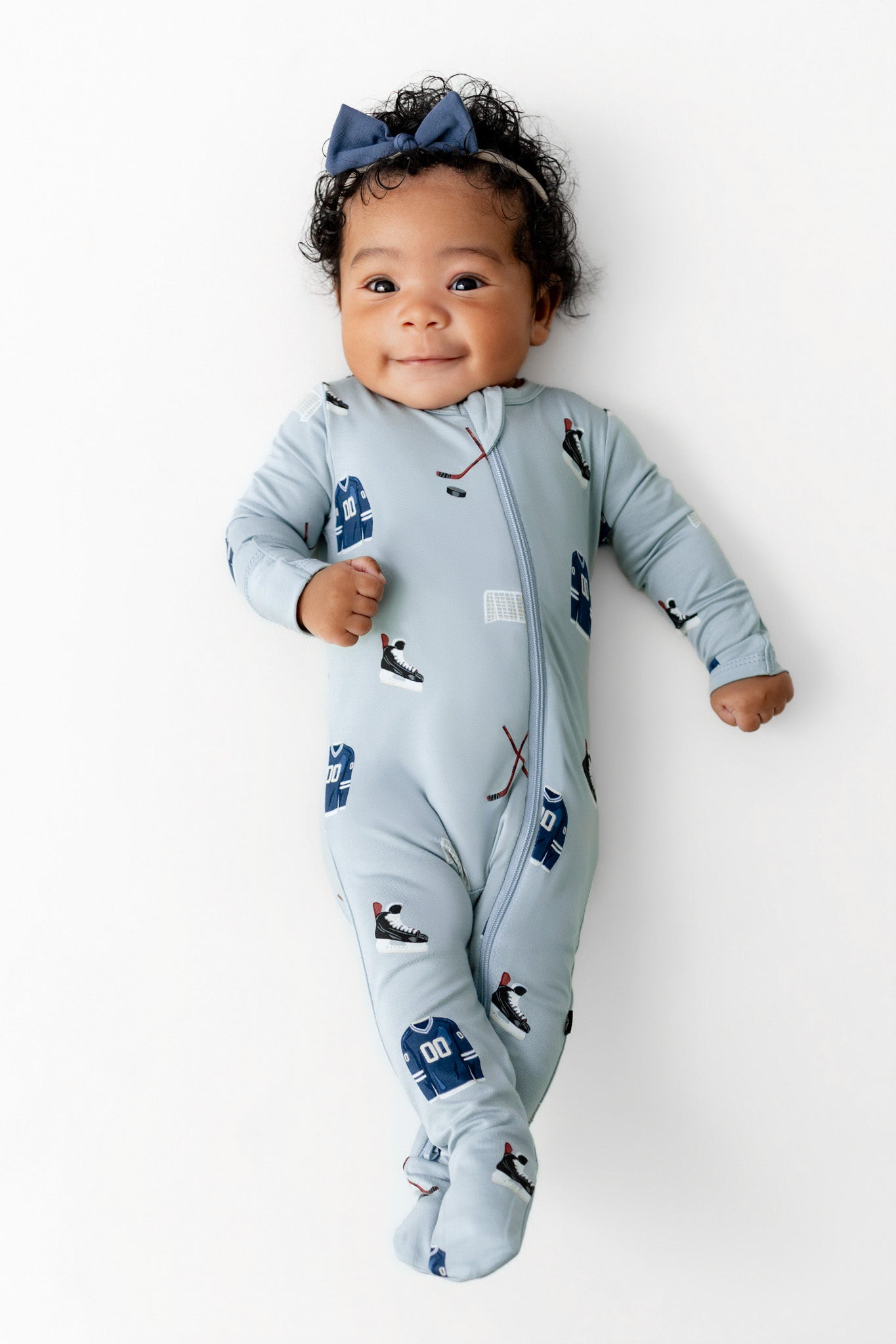Baby wearing Zippered Footie in Vintage Hockey print and blue bow headband on a white background