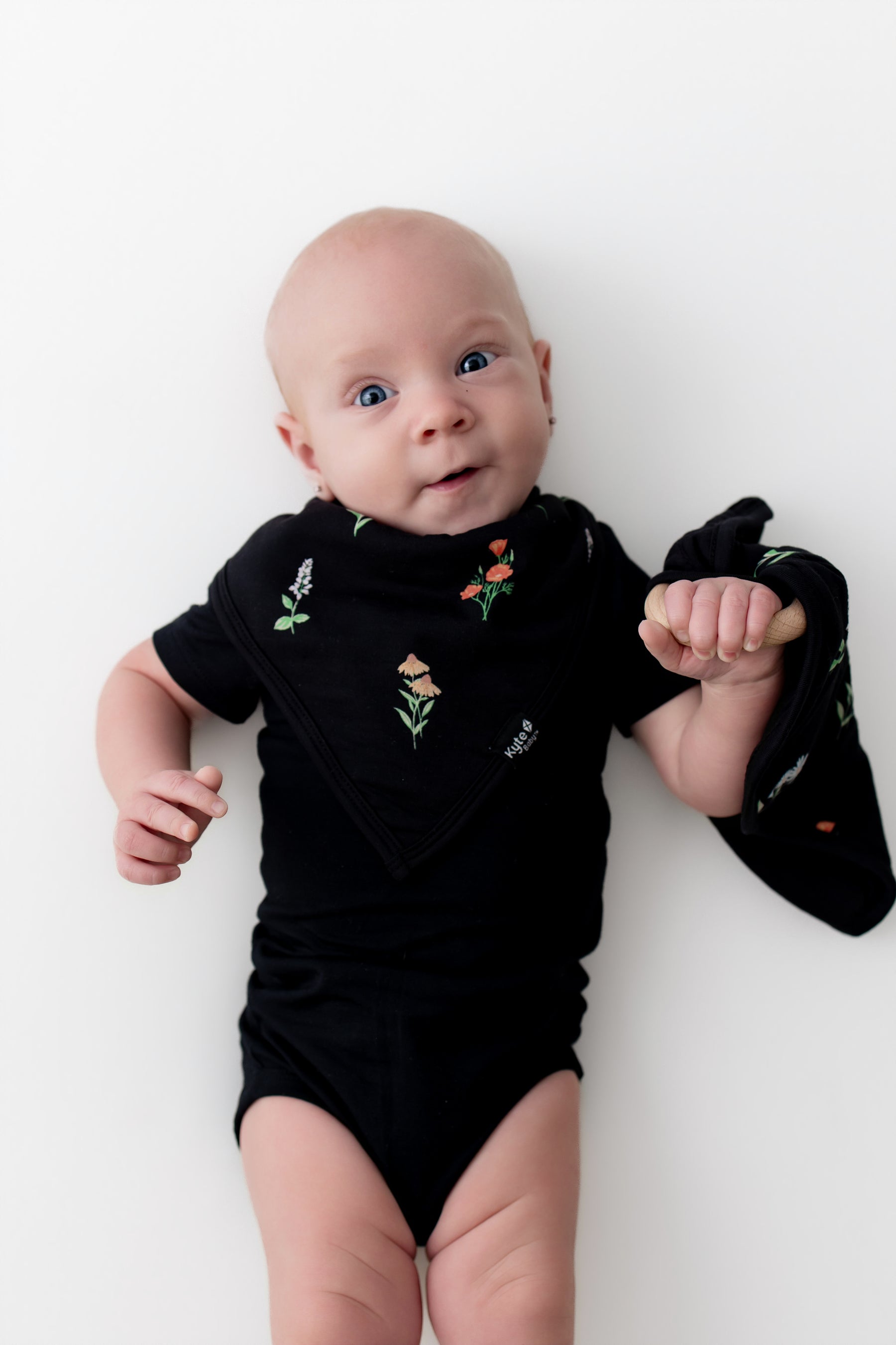 Baby with Lovey in Midnight Wildflower with Removable Wooden Teething Ring wearing a black bodysuit and midnight wildflower bib on a white background