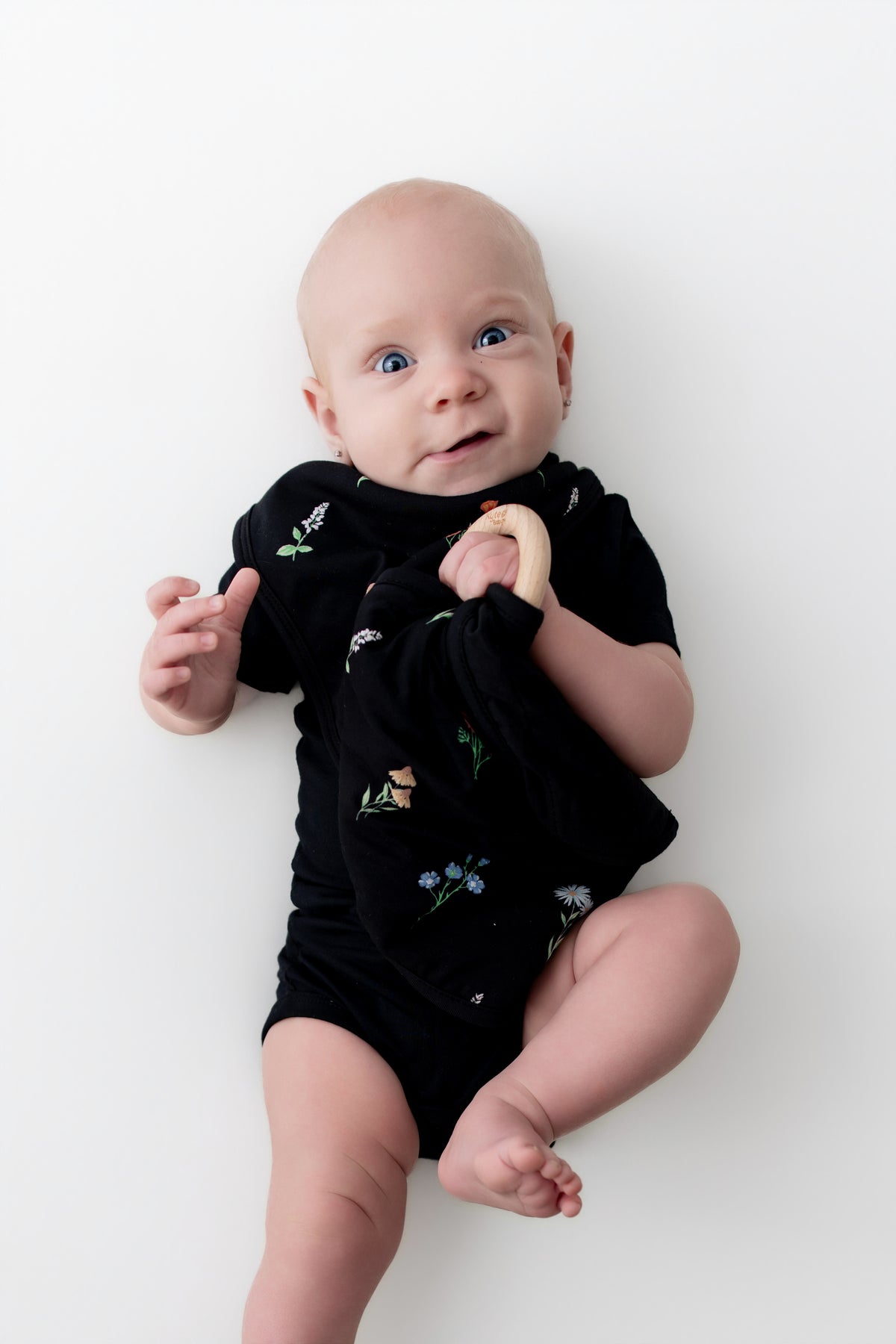 Baby holding Lovey in Midnight Wildflower with Removable Wooden Teething Ring wearing a black bodysuit and midnight wildflower bib on a white background