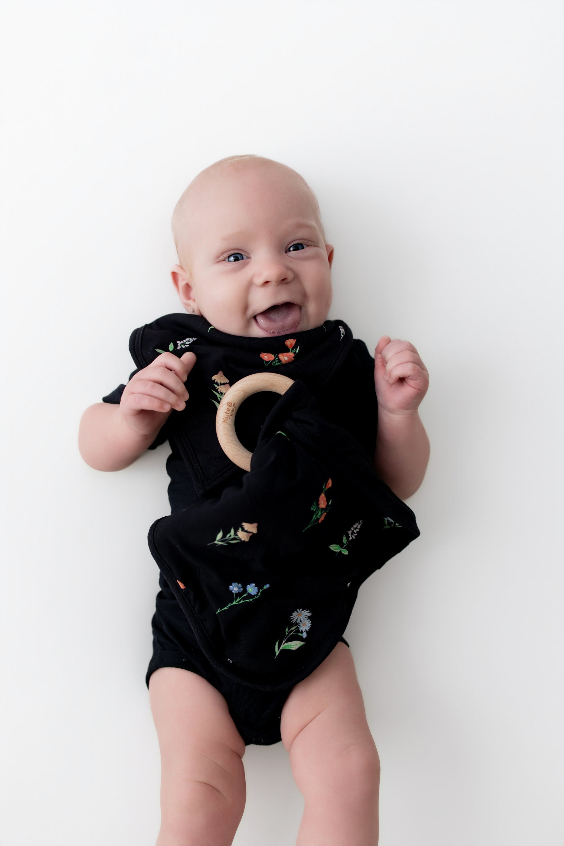 Baby holding Lovey in Midnight Wildflower with Removable Wooden Teething Ring wearing a black bodysuit and bib in midnight wildflower print on a white background