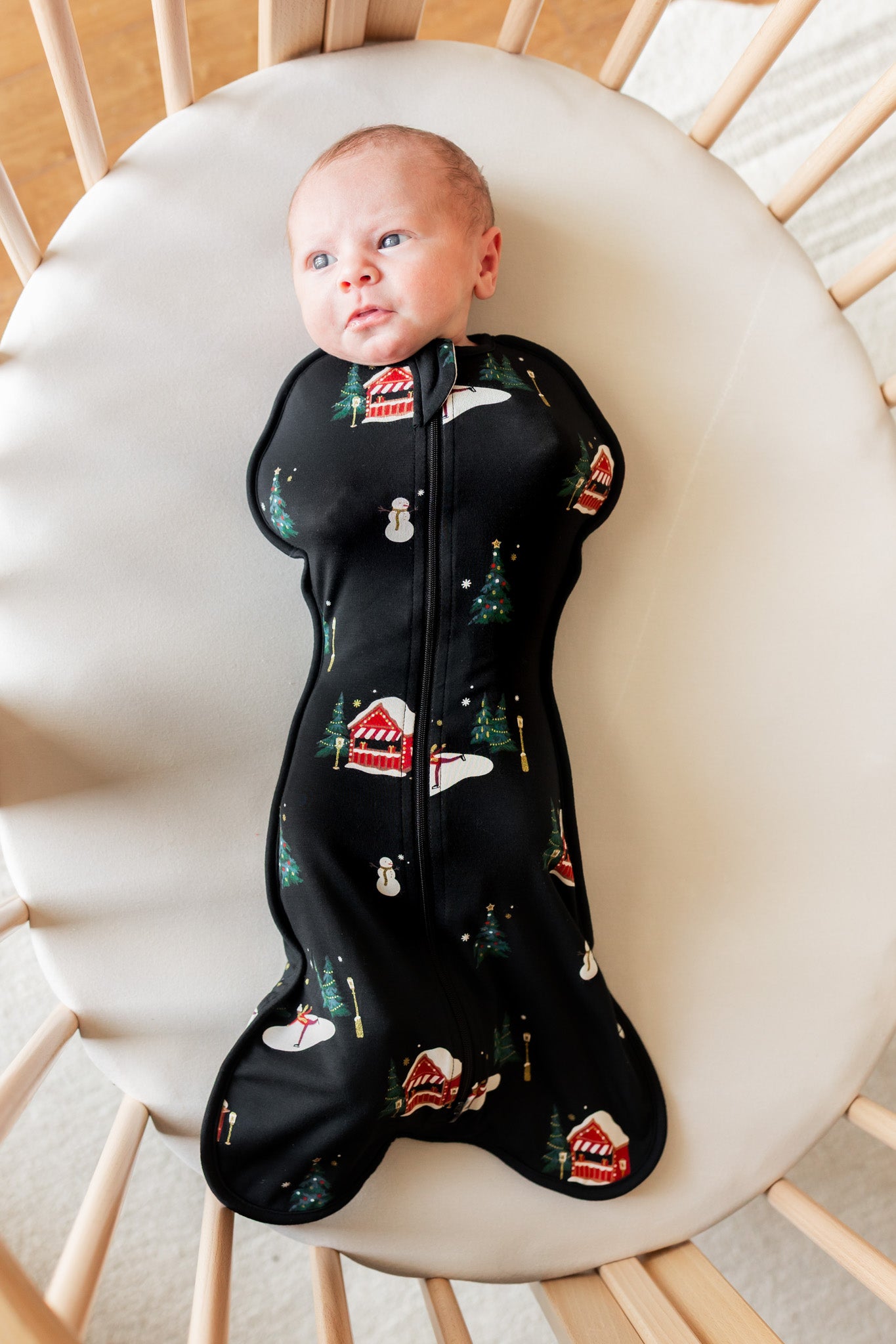 A baby lying on their back in a wooden crib, wearing a black sleep sack with a Christmas print of snowmen, trees, and houses.