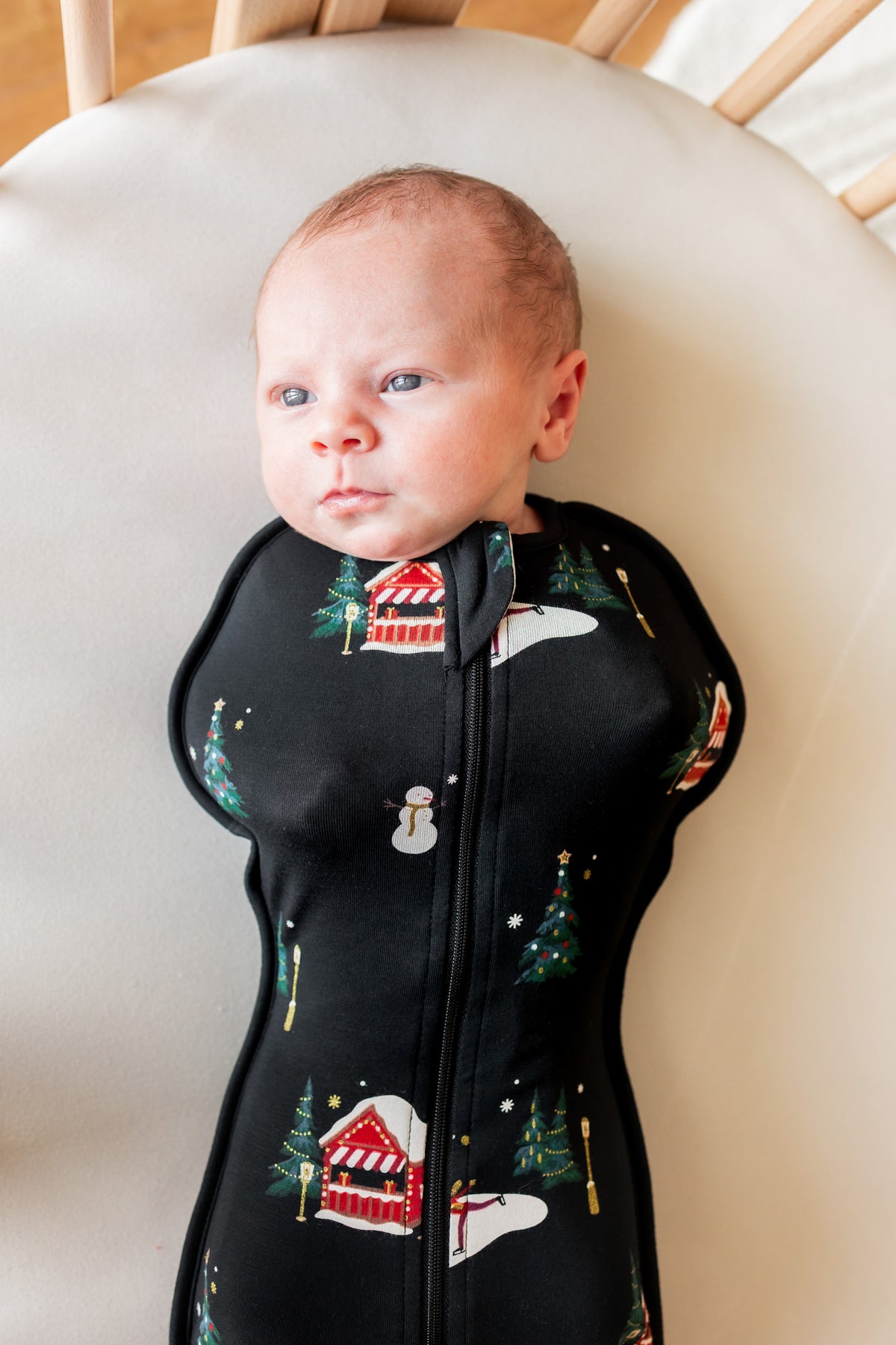 A baby wearing a black zip-up sleeper with a Christmas print of snowmen, trees, and houses, lying on a beige cushion.