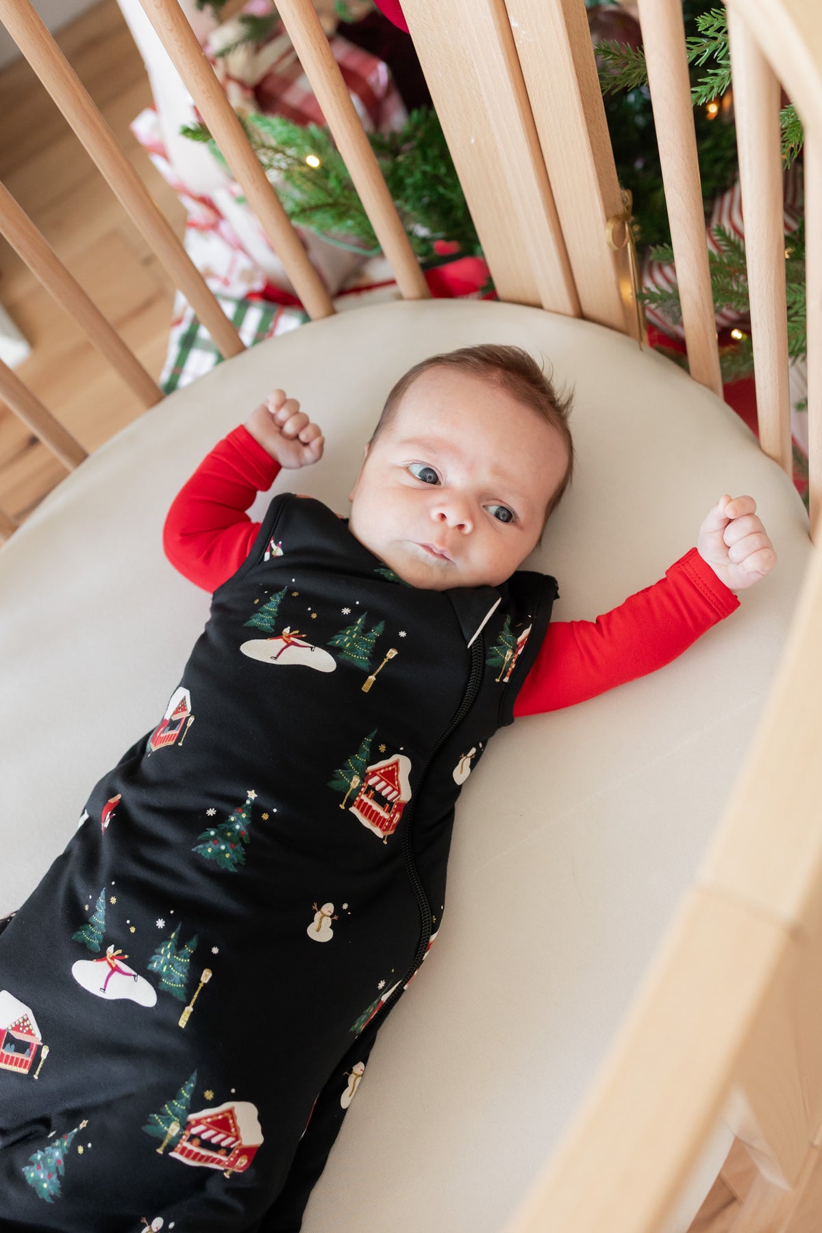 A baby lying on their back in a wooden crib, wearing a black Christmas-themed sleeping bag with a red long-sleeve shirt underneath. The sleeping bag features a pattern of snowman, Christmas tree, and snowman house motifs.