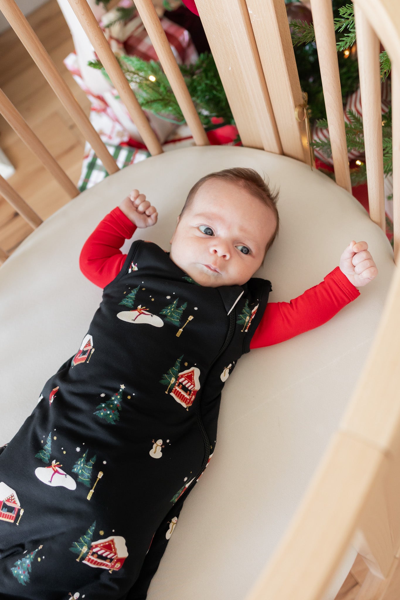 A baby lying on their back in a wooden crib, wearing a black Christmas-themed sleeping bag with a red long-sleeve shirt underneath. The sleeping bag features a pattern of snowman, Christmas tree, and snowman house motifs.
