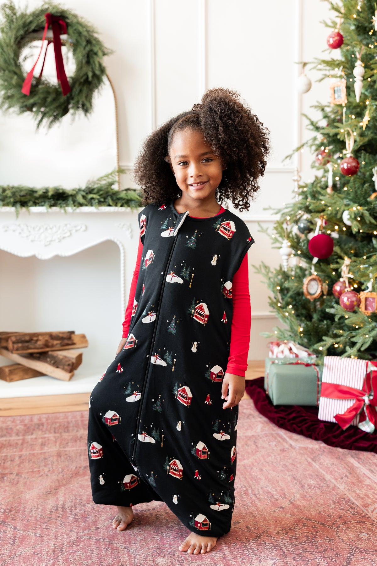 A young girl with curly hair stands barefoot in a holiday-themed room, wearing a black zip-up sleep sack with a Christmas print over a red long-sleeve shirt. A decorated Christmas tree, fireplace, and wrapped gifts are visible in the background.
