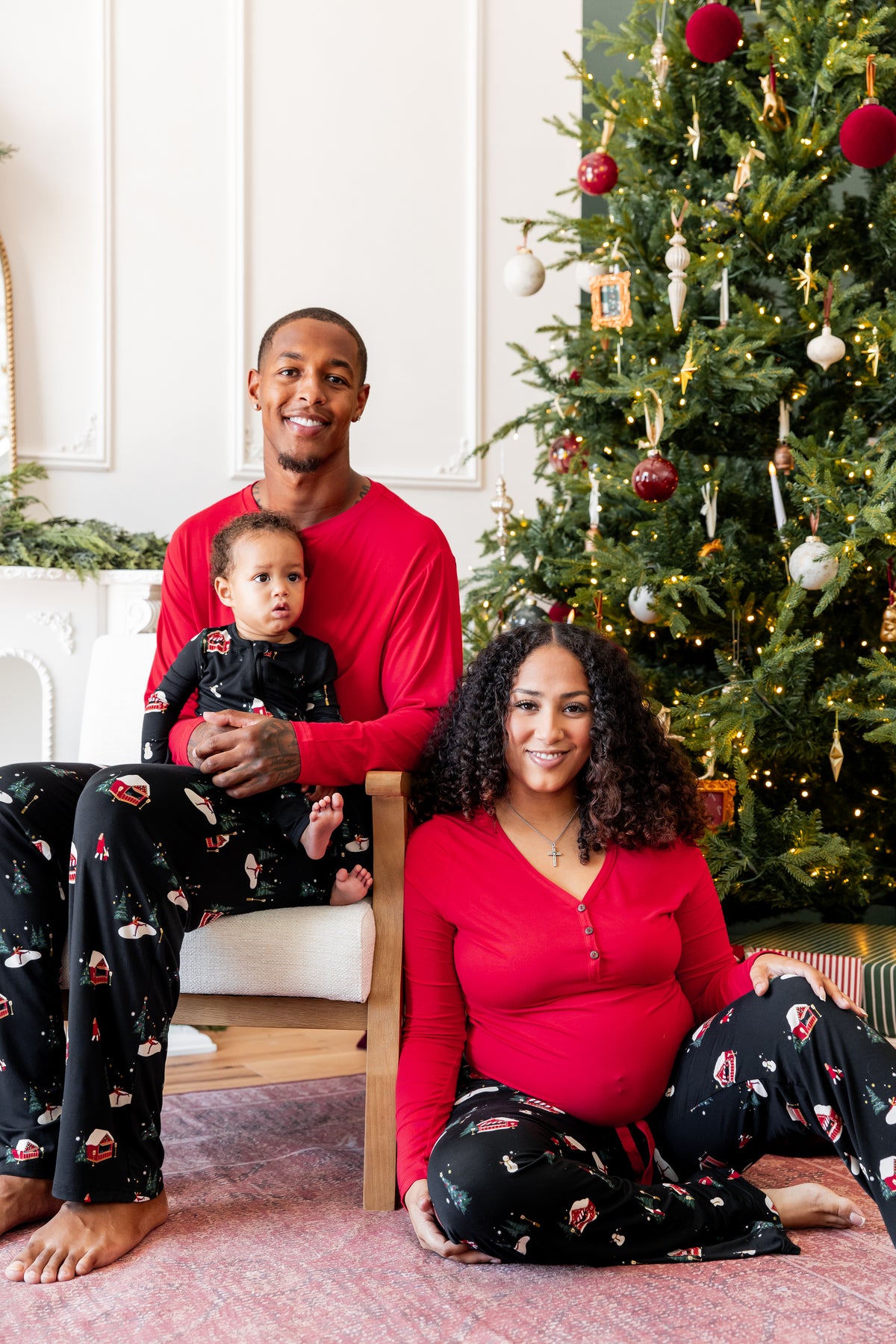 A family of three wearing matching Christmas-themed pajamas, with a pregnant woman sitting on the floor, a man holding a baby on a chair, and a decorated Christmas tree in the background.