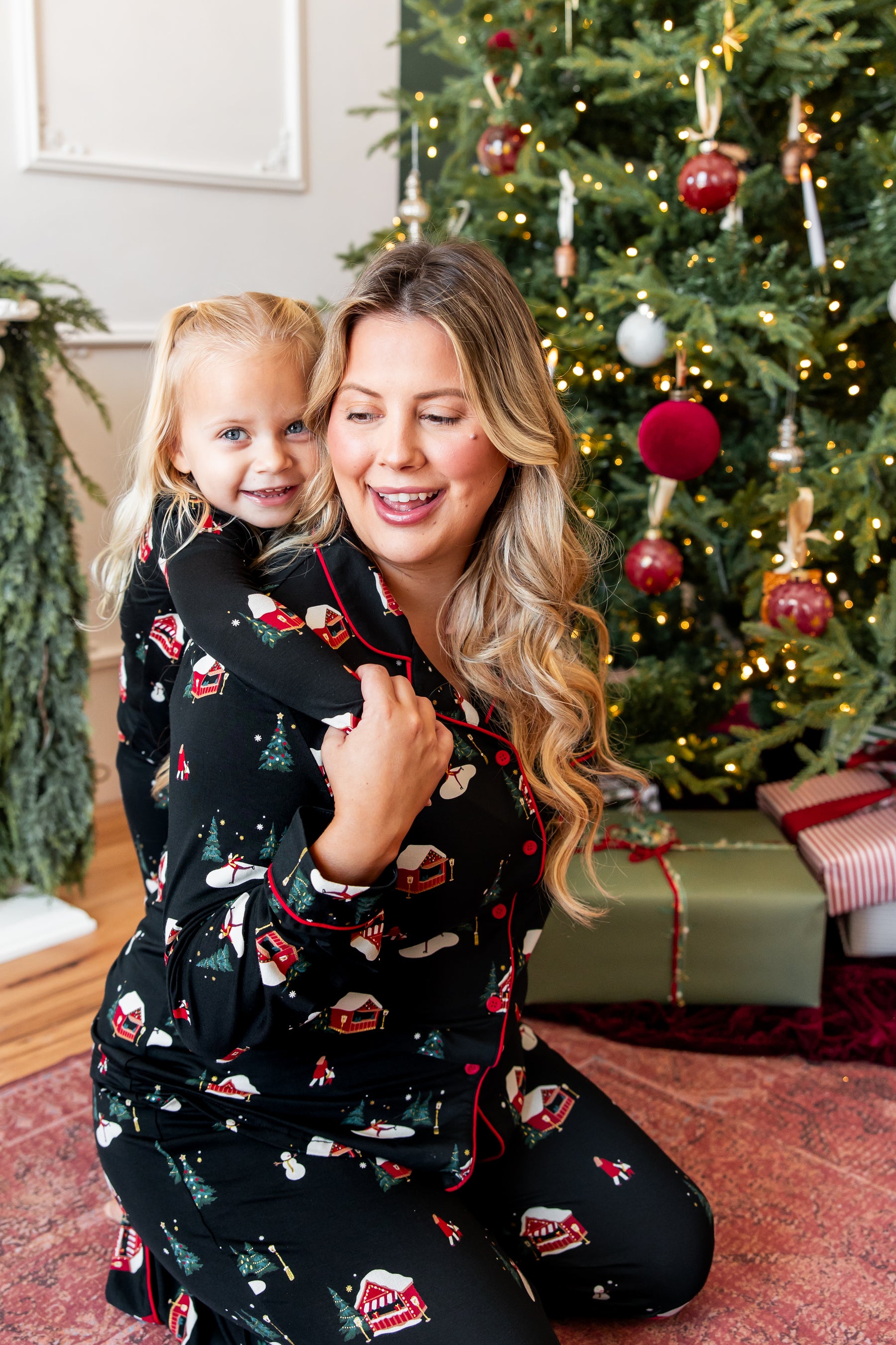 A woman and a young girl wearing matching black Christmas-themed pajamas with red trim and holiday motifs, kneeling in front of a decorated Christmas tree in a cozy indoor setting.