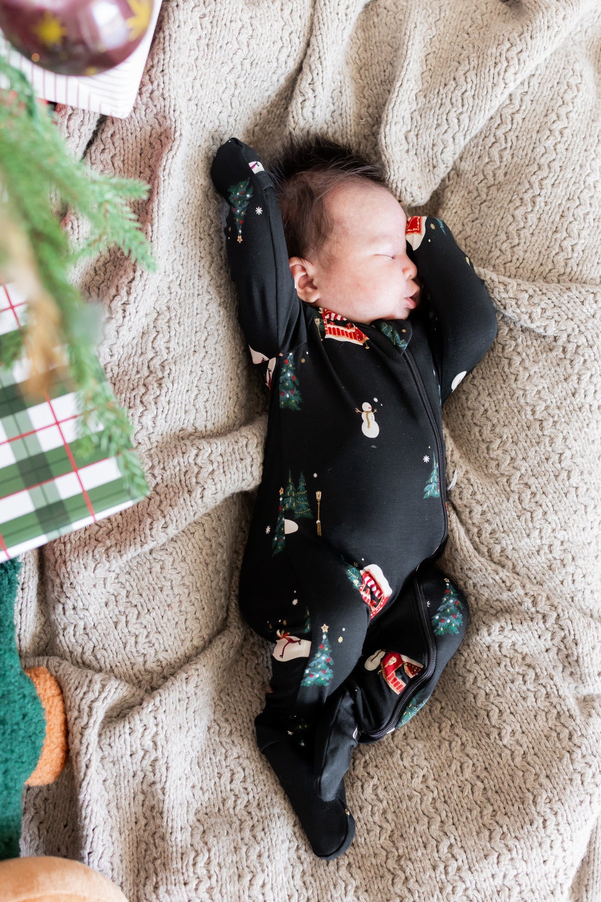 A newborn baby sleeping soundly on their back, wearing a black footed sleeper with a festive Christmas print of snowmen, trees, and ornaments, lying on a cozy beige knit blanket with holiday decorations in the background.
