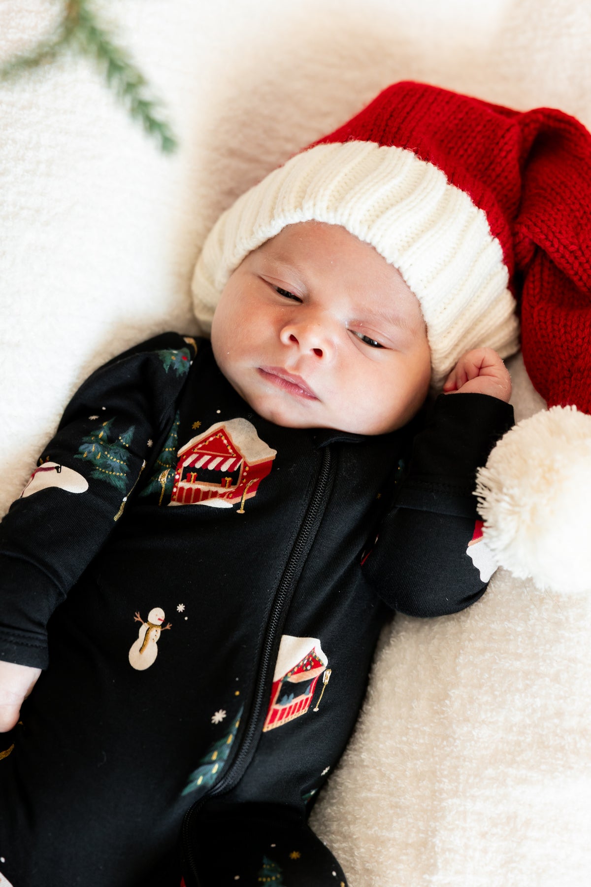 A sleeping newborn baby wearing a black Christmas-themed zip-up onesie with festive house and snowman prints, paired with a red and white knit Santa hat, lying on a soft cream blanket.