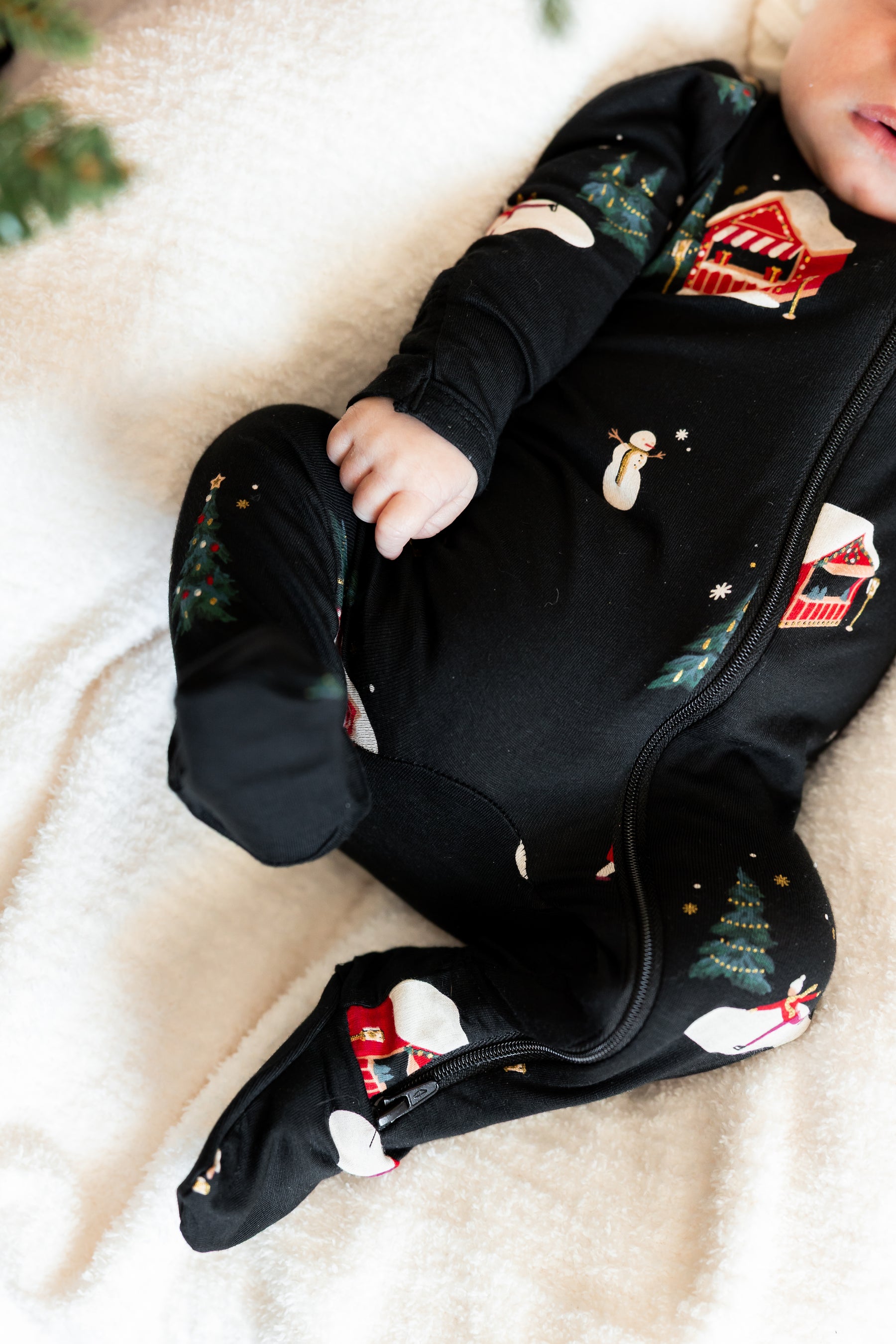 A baby lying on a soft cream blanket wearing a black footed onesie with a Christmas print featuring snowmen, trees, and red holiday motifs.