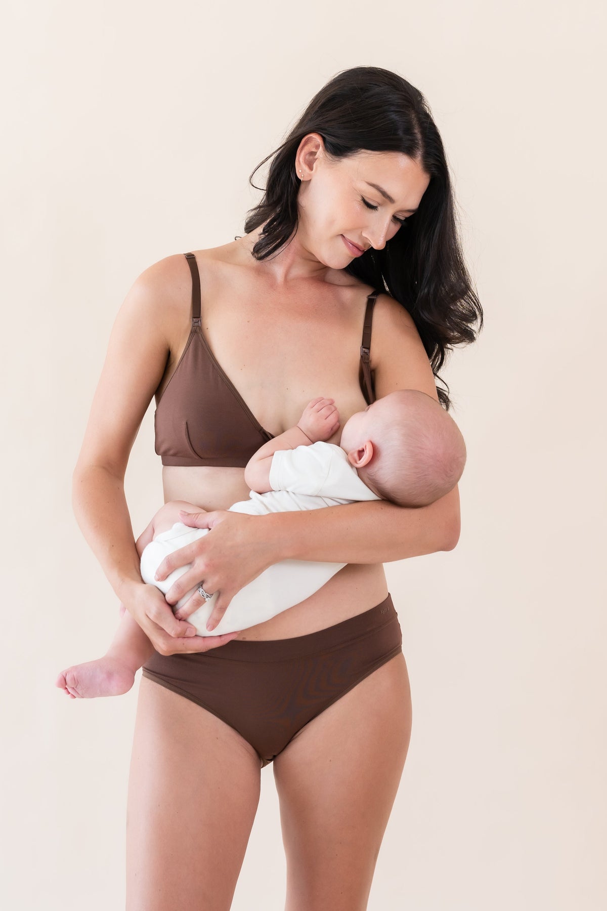 Woman in brown bra and underwear holding a baby against a beige background