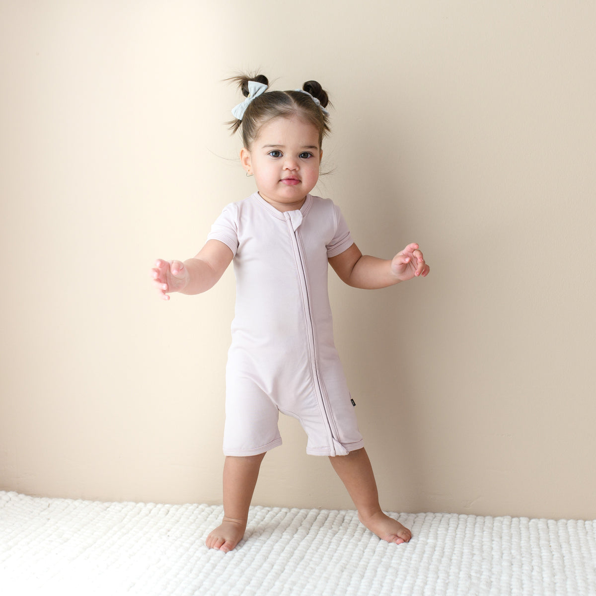 Toddler standing infront of beige wall while wearing dual Zipper Shortall Romper in Blush 