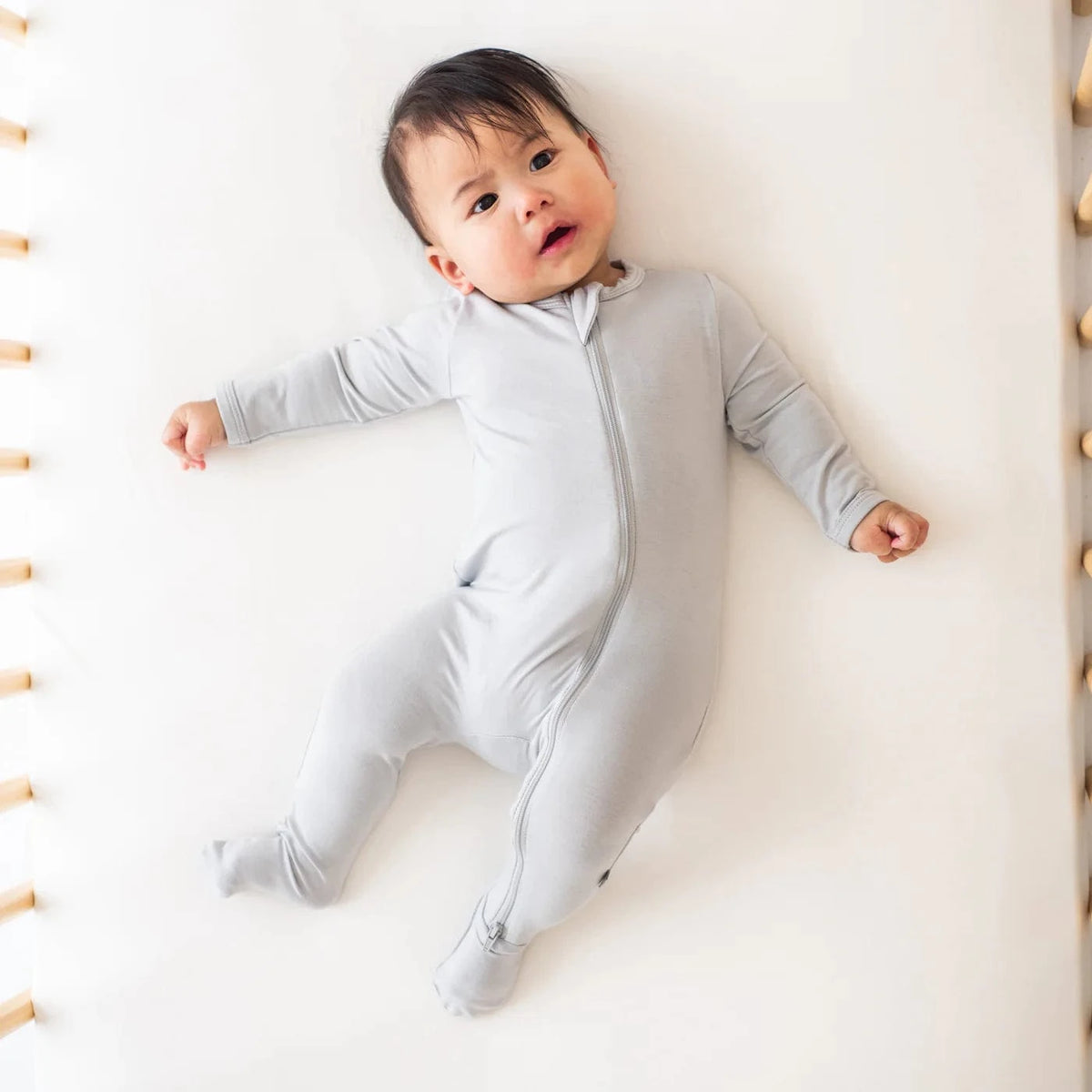Infant laying in a crib on a light neutral crib sheet wearing the Zippered Footie in Storm