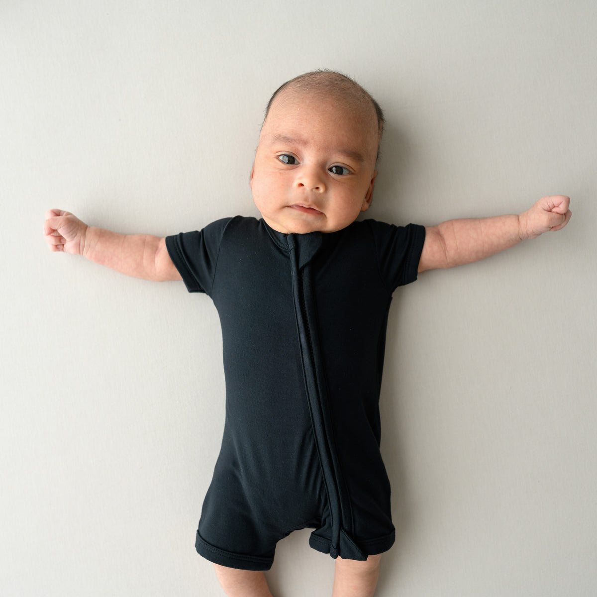 Infant lying on an oat surface while wearing a Zippered Shortall Romper in Midnight