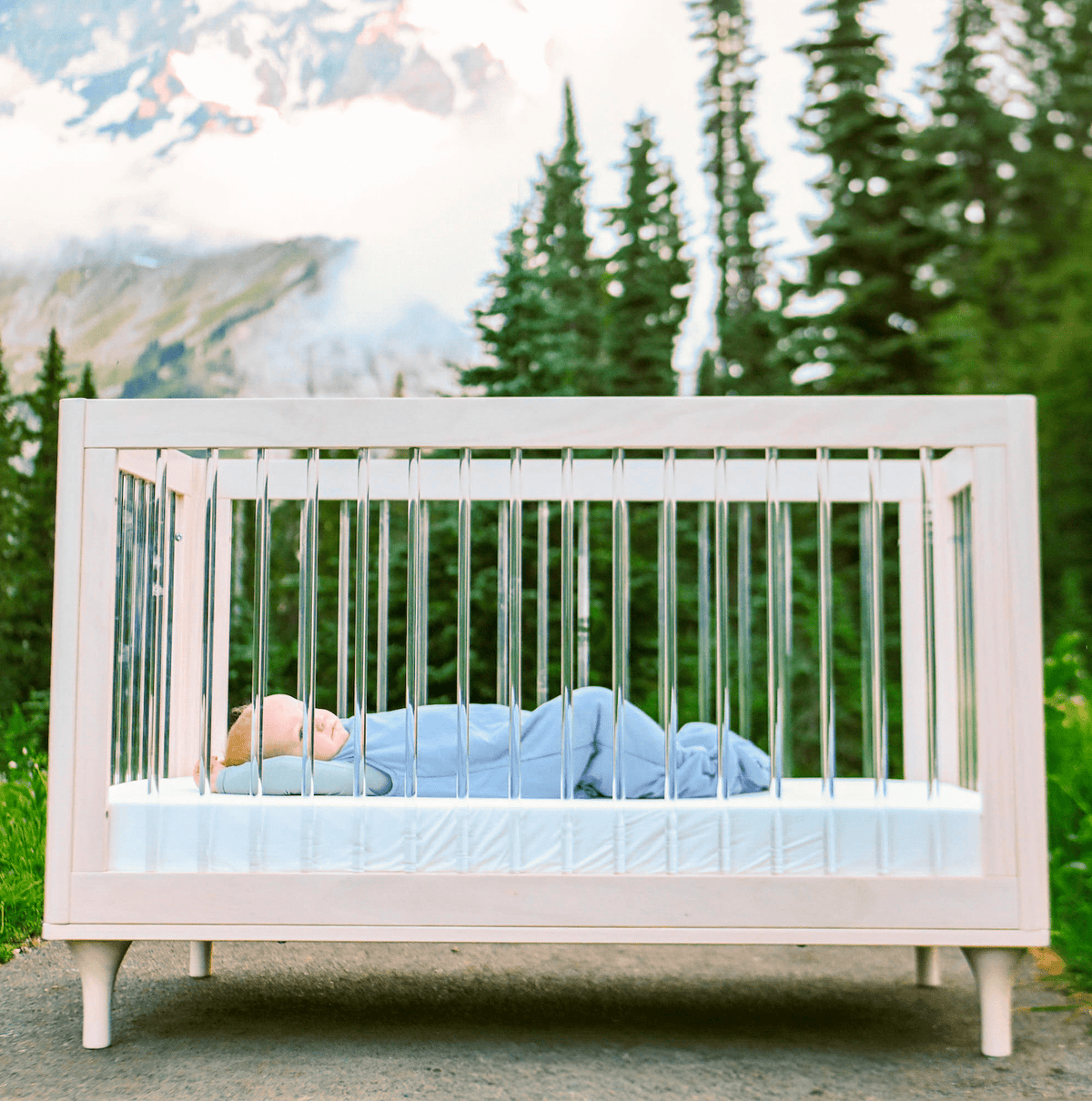 Baby in Slate Sleep Bag laying in a crib with a forest in the background 