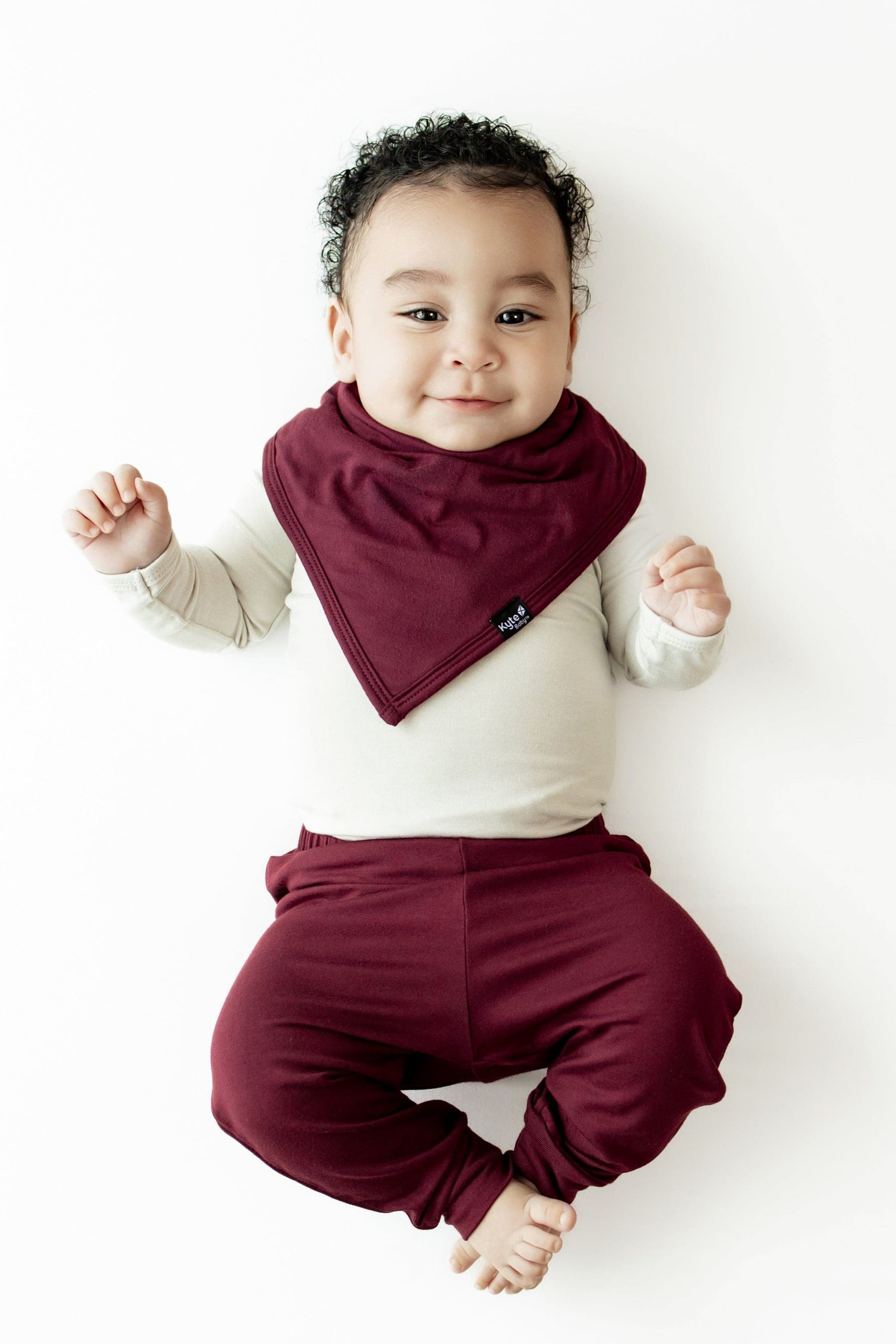A baby lying on their back wearing a burgundy triangle bib over a cream long-sleeve top and matching burgundy pants, photographed against a clean white background.