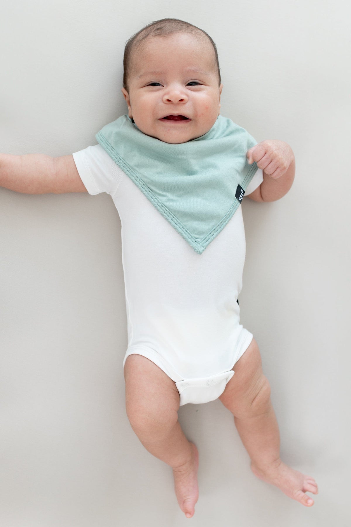 Baby laying on a Crib Sheet in Oat while wearing a  Bib in Sage