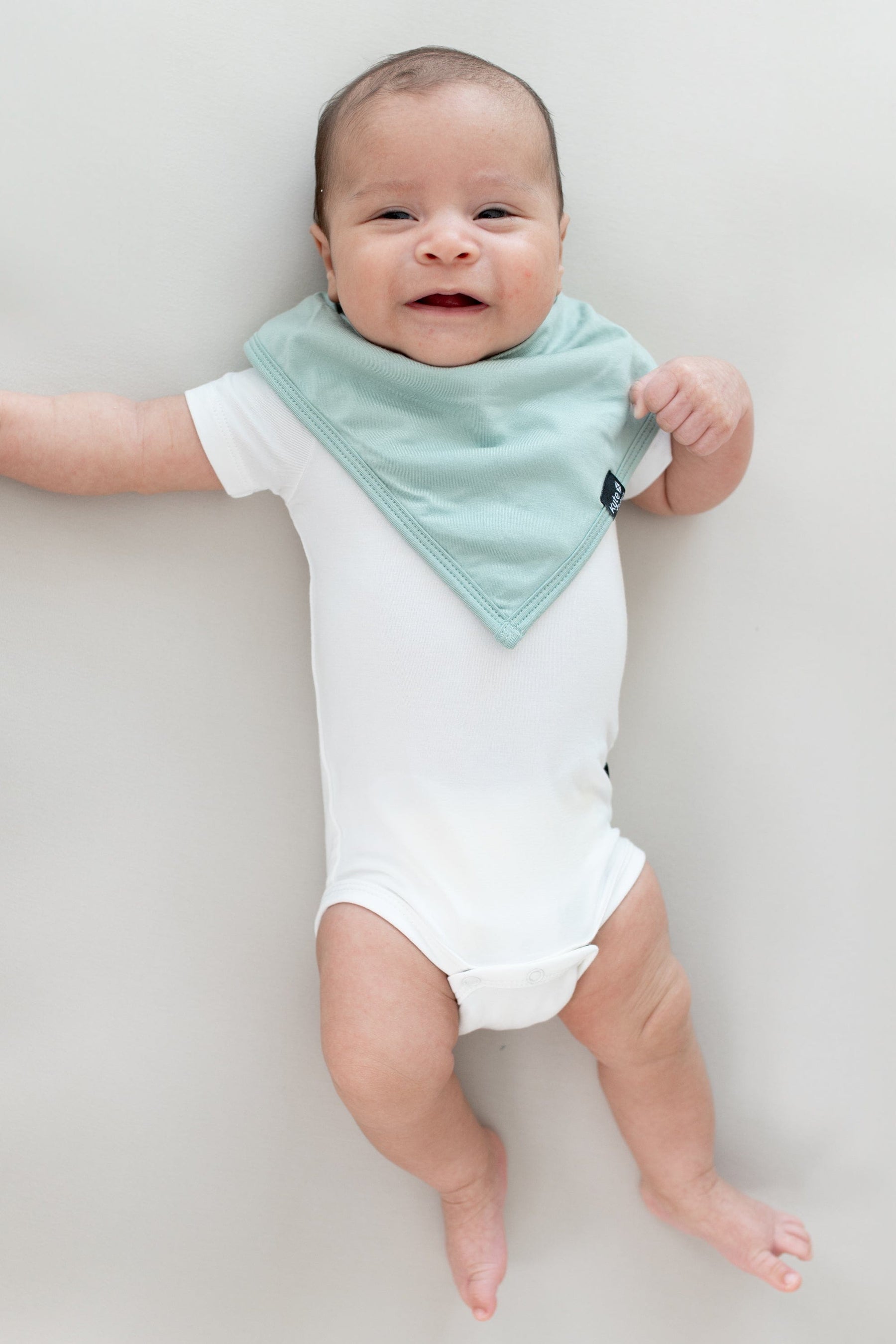 Baby laying on a Crib Sheet in Oat while wearing a  Bib in Sage