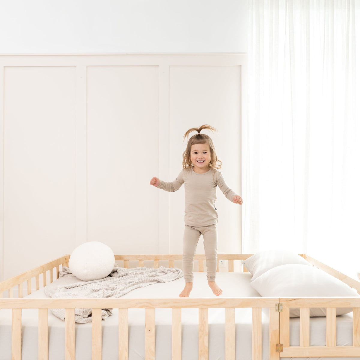 Toddler jumping on bed made with fitted sheet wearing two piece Kyte Baby Long Sleeve Pajamas in Almond