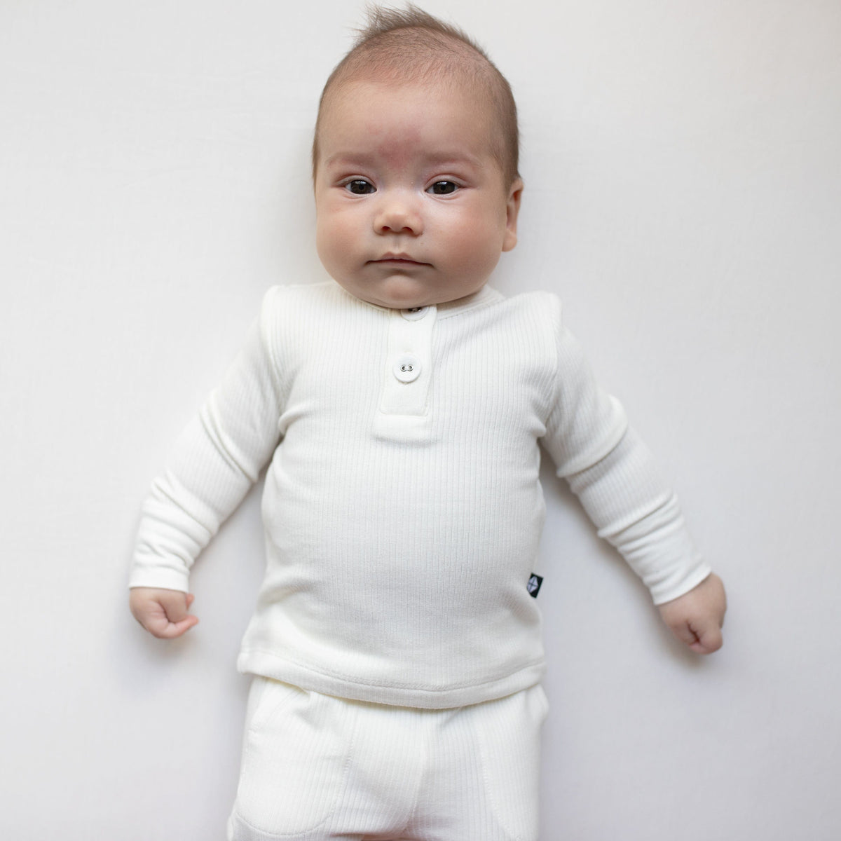 Infant laying on white surface wearing Kyte Baby Bamboo Ribbed Infant Henley Set in Cloud white