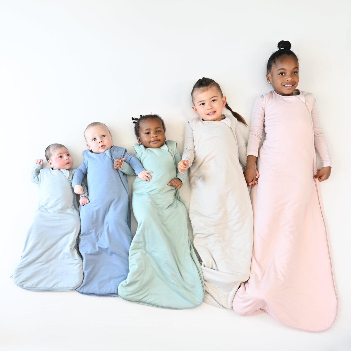 Five child models laying side by side from smallest to largest wearing 1.0 TOG Kyte Baby Sleep Bags in core colors  