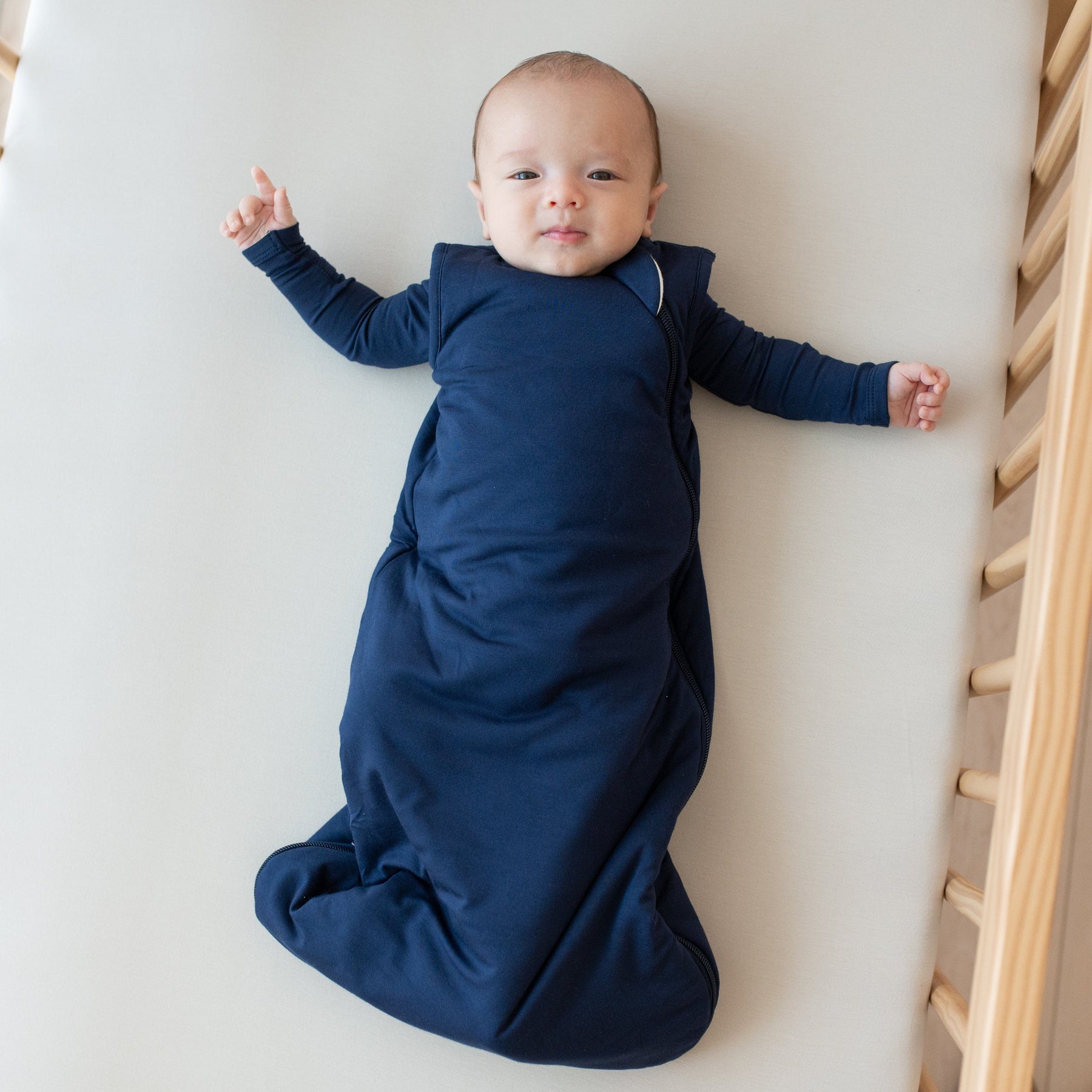 Baby laying in a crib while wearing a Kyte Baby Sleep Bag in Navy 1.0 and matching bodysuit underneath 