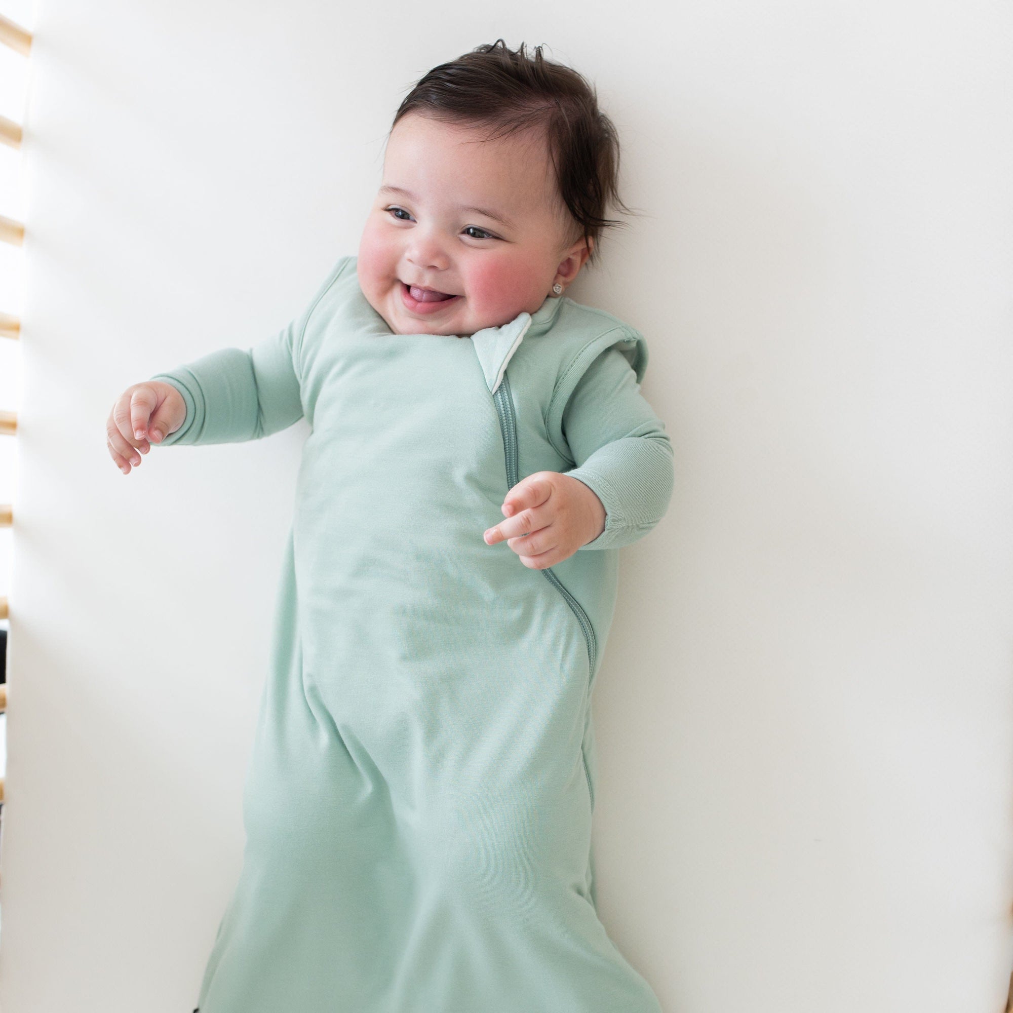 Smiling baby looking to the left and wearing a Kyte Baby Sleep Bag in Sage 1.0