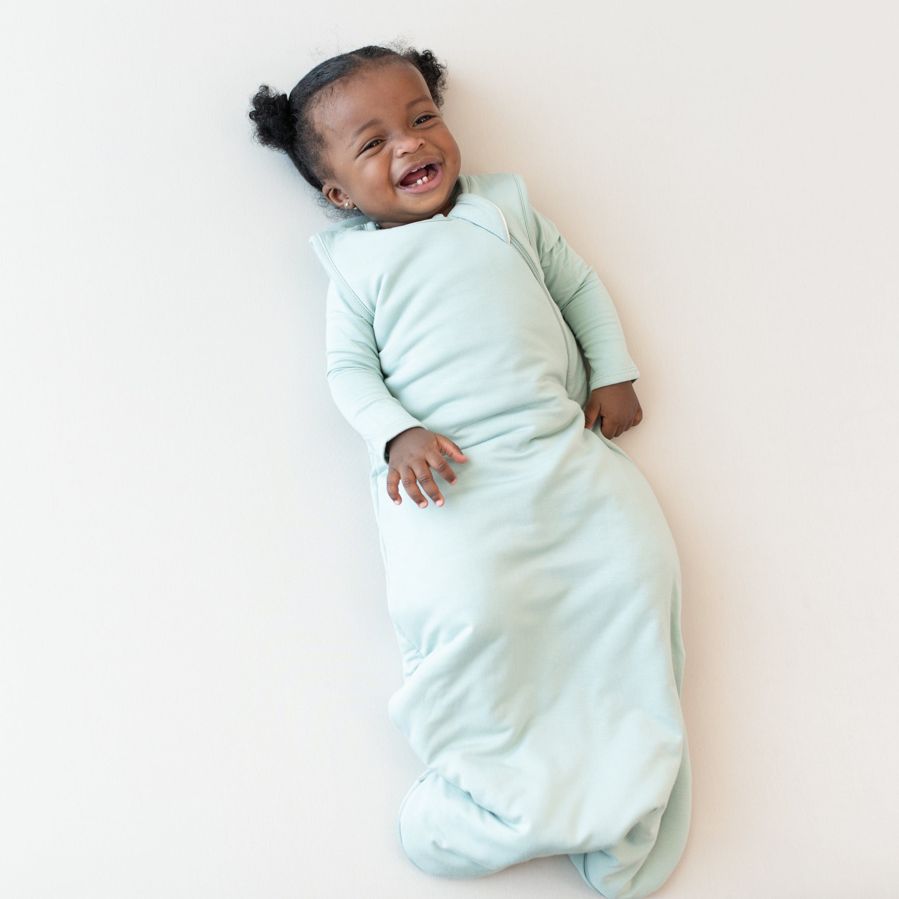 Smiling baby laying on an oat background while wearing a Sleep Bag in Sage 1.0 with a long sleeve Sage bodysuit underneath