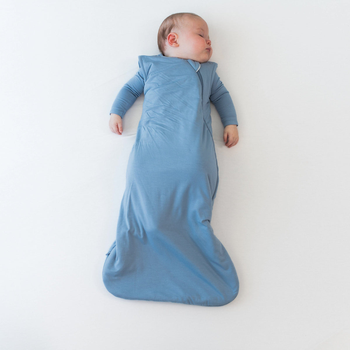 Baby sleeping on a flat off-white surface in the 2.5 Sleep Bag in Slate 