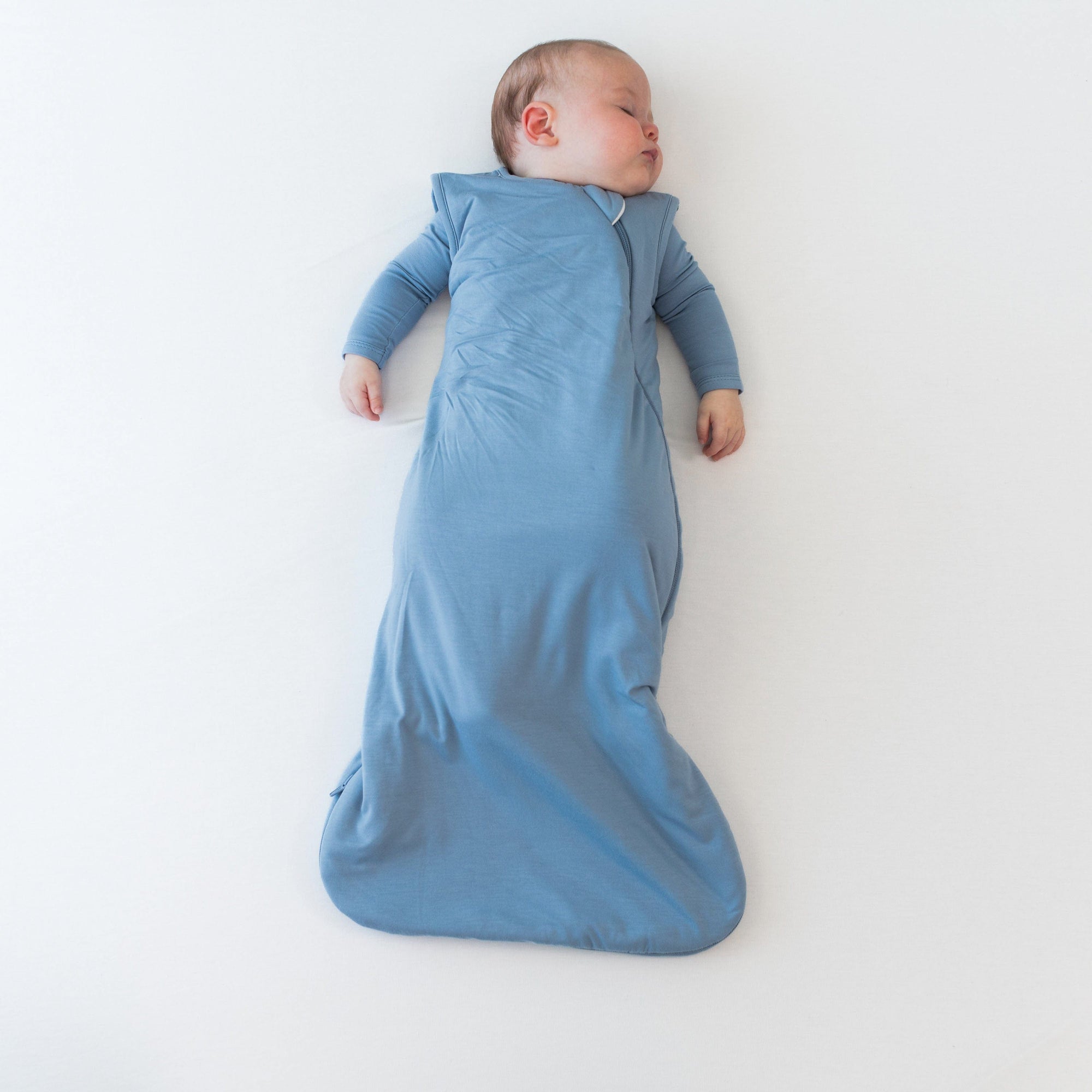 Baby sleeping on a flat off-white surface in the 2.5 Sleep Bag in Slate 