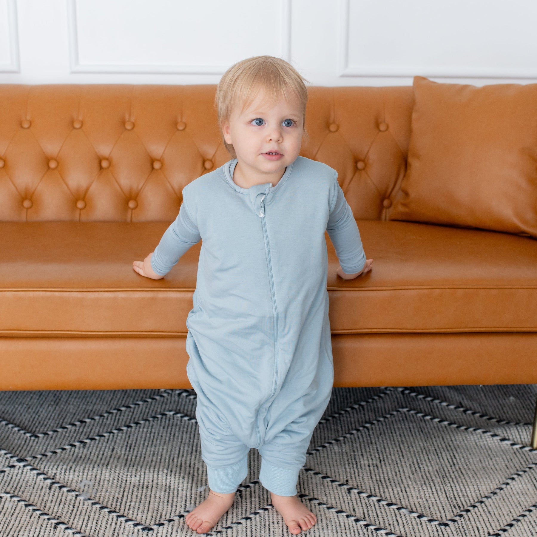 Toddler standing in front of a couch wearing Kyte Baby Sleep Bag Walker in Fog 1.0